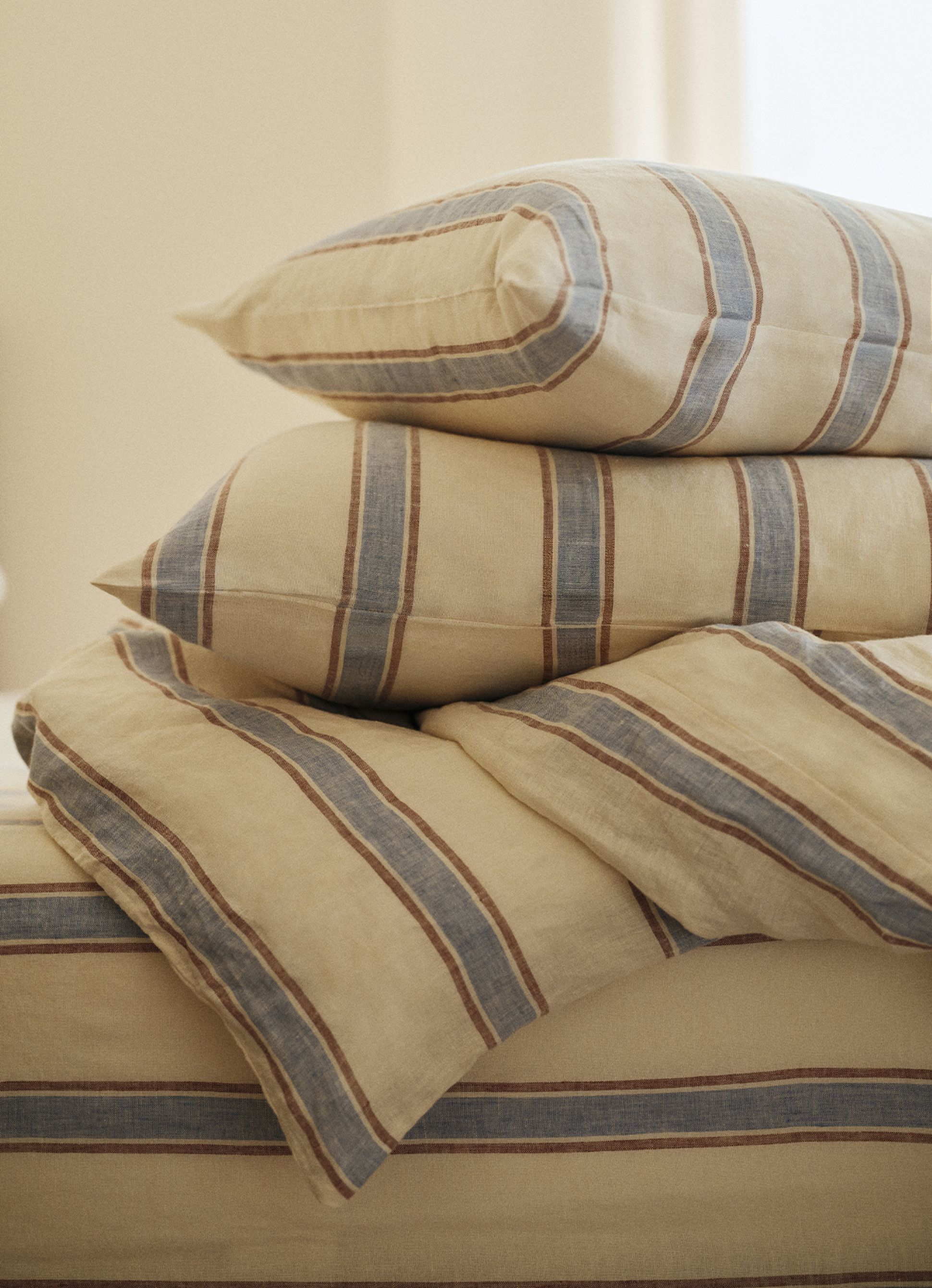 Une pile de coussins rayés et de linge de lit plié, dans des tons doux de beige, de bleu et de marron, créant une ambiance chaleureuse et accueillante.