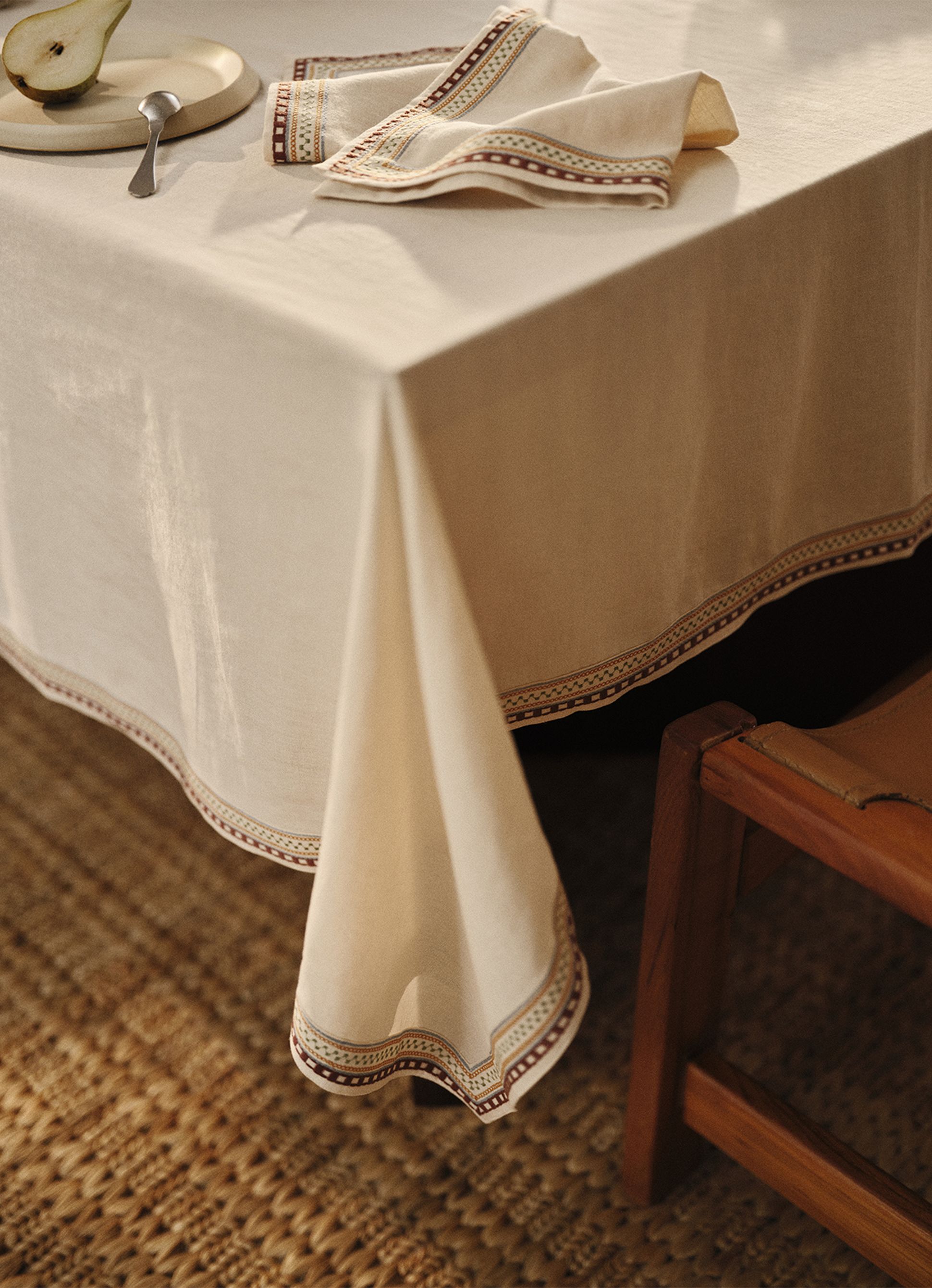 Une nappe crème à bordure à motifs recouvre une table sur laquelle sont posés une assiette, une demi-poire et une serviette, à côté d'une chaise en bois posée sur une natte tressée.