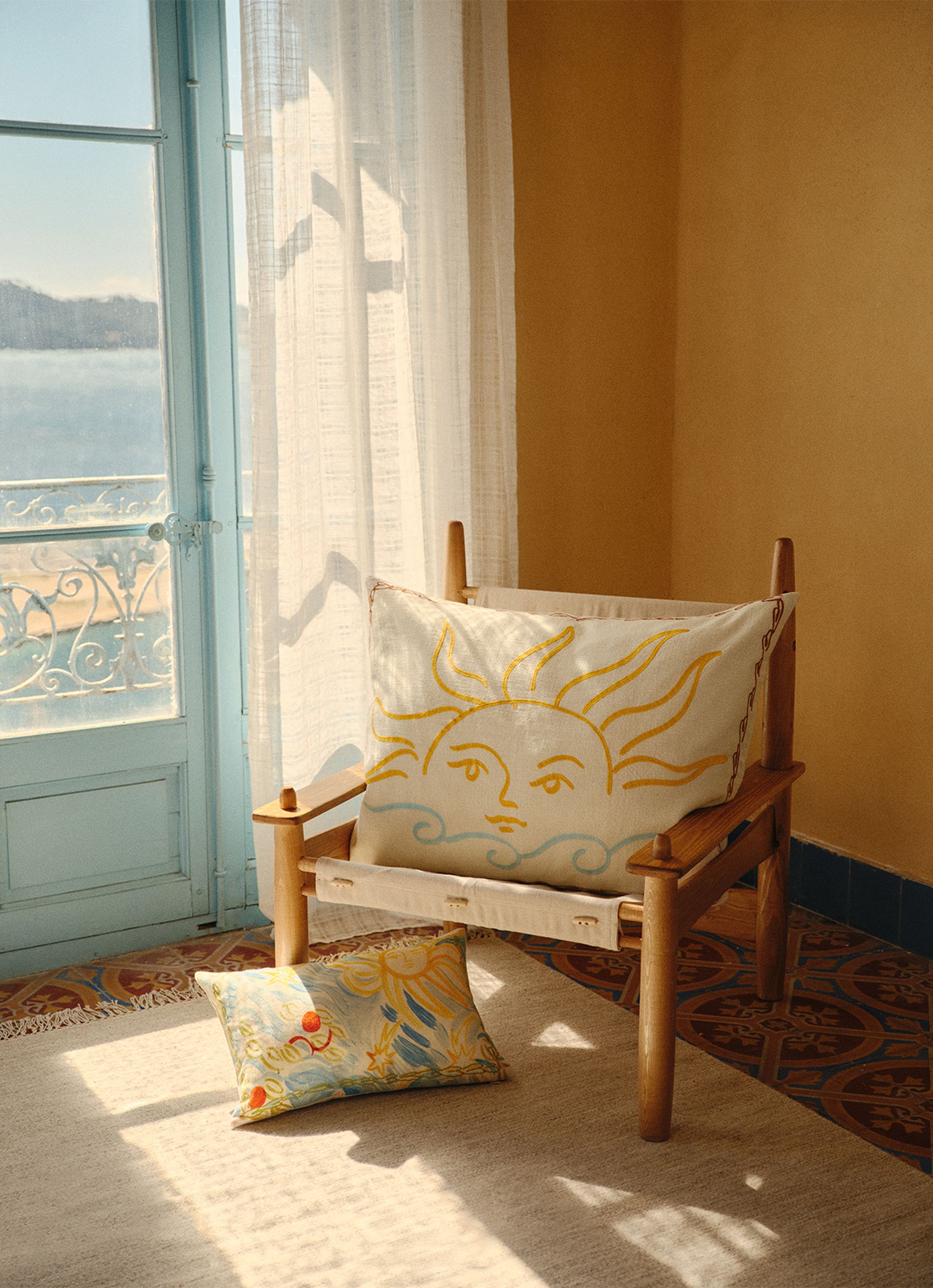 Une chaise en bois avec un coussin orné d'un motif solaire, placée près d'une fenêtre ensoleillée, avec des rideaux vaporeux et une vue sur un lac. Un coussin à motifs repose à proximité, sur le sol.