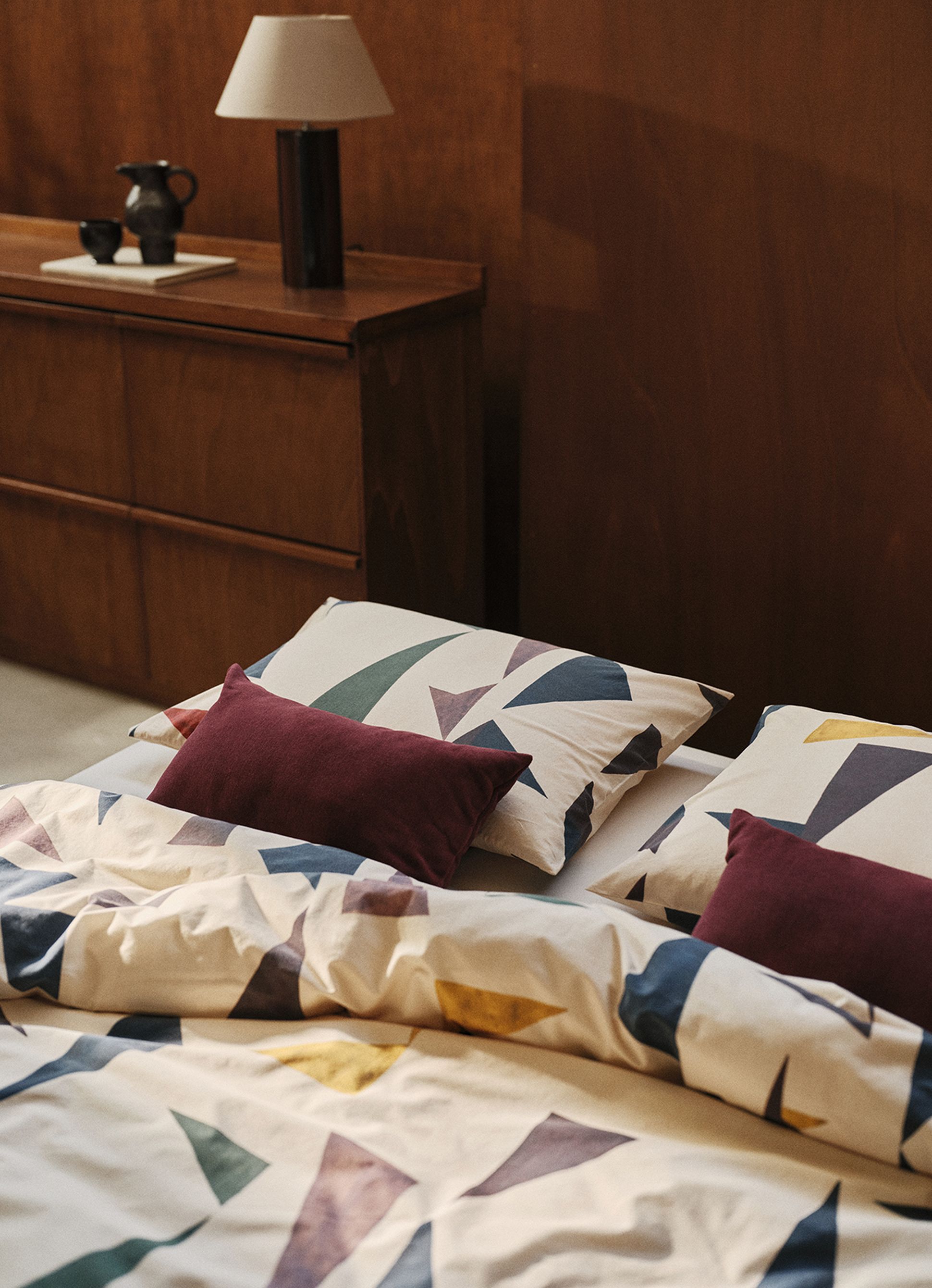 Une parure de lit aux motifs géométriques, agrémentée de coussins rouges, sur un lit. À côté se trouve une table de chevet en bois sur laquelle sont posés une lampe et un objet en poterie.