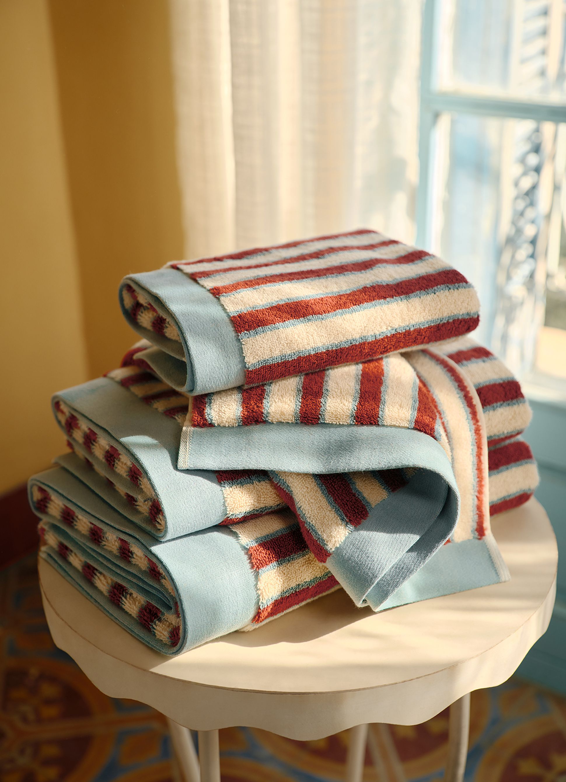 Une pile de serviettes pliées à rayures rouges, bleues et blanches posée sur une petite table ronde, éclairée par la lumière du soleil près d'une fenêtre.