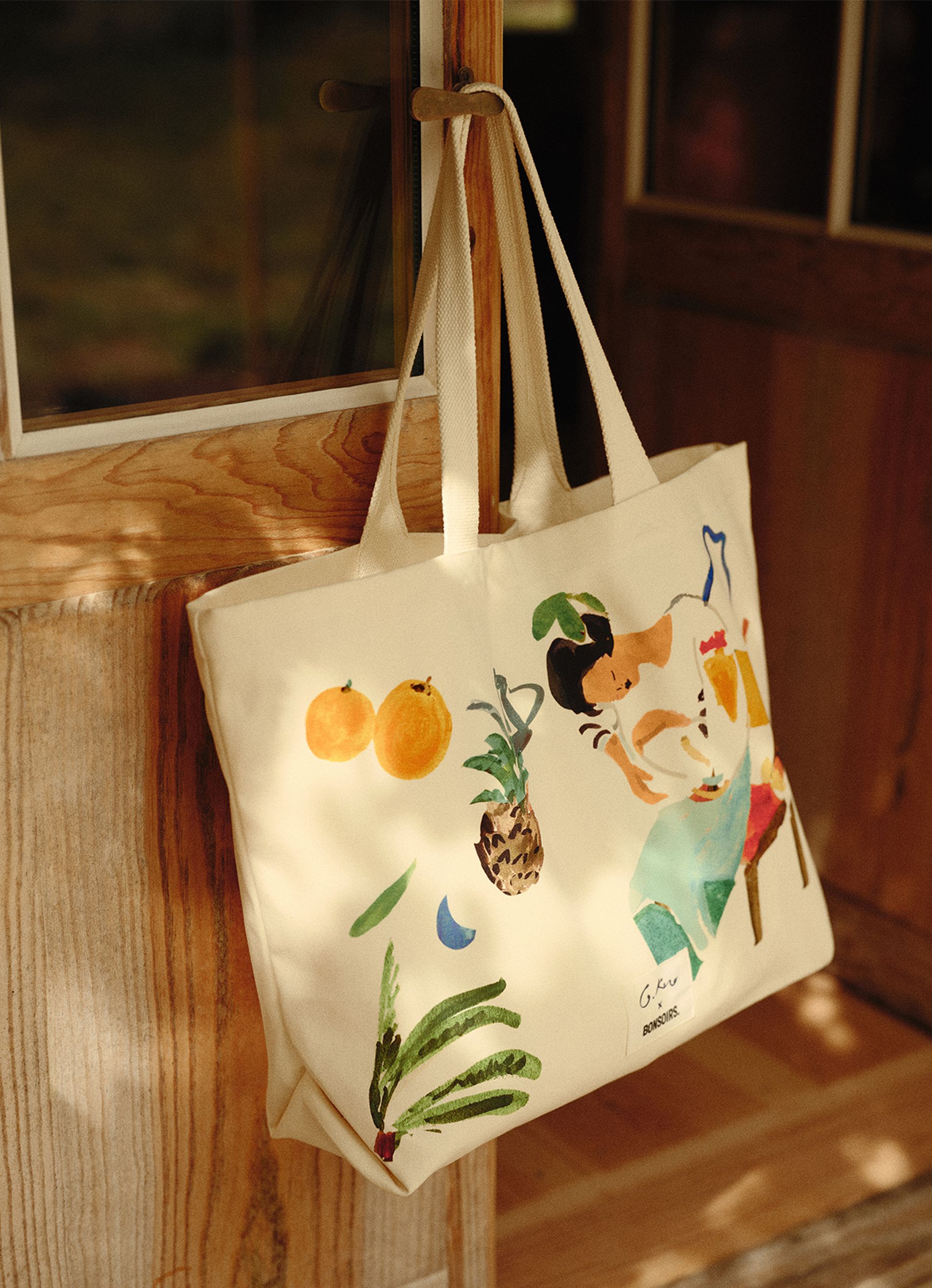 Sac fourre-tout en toile orné d'une illustration colorée représentant des fruits et une femme, suspendu à une poignée de porte sous un éclairage chaleureux.