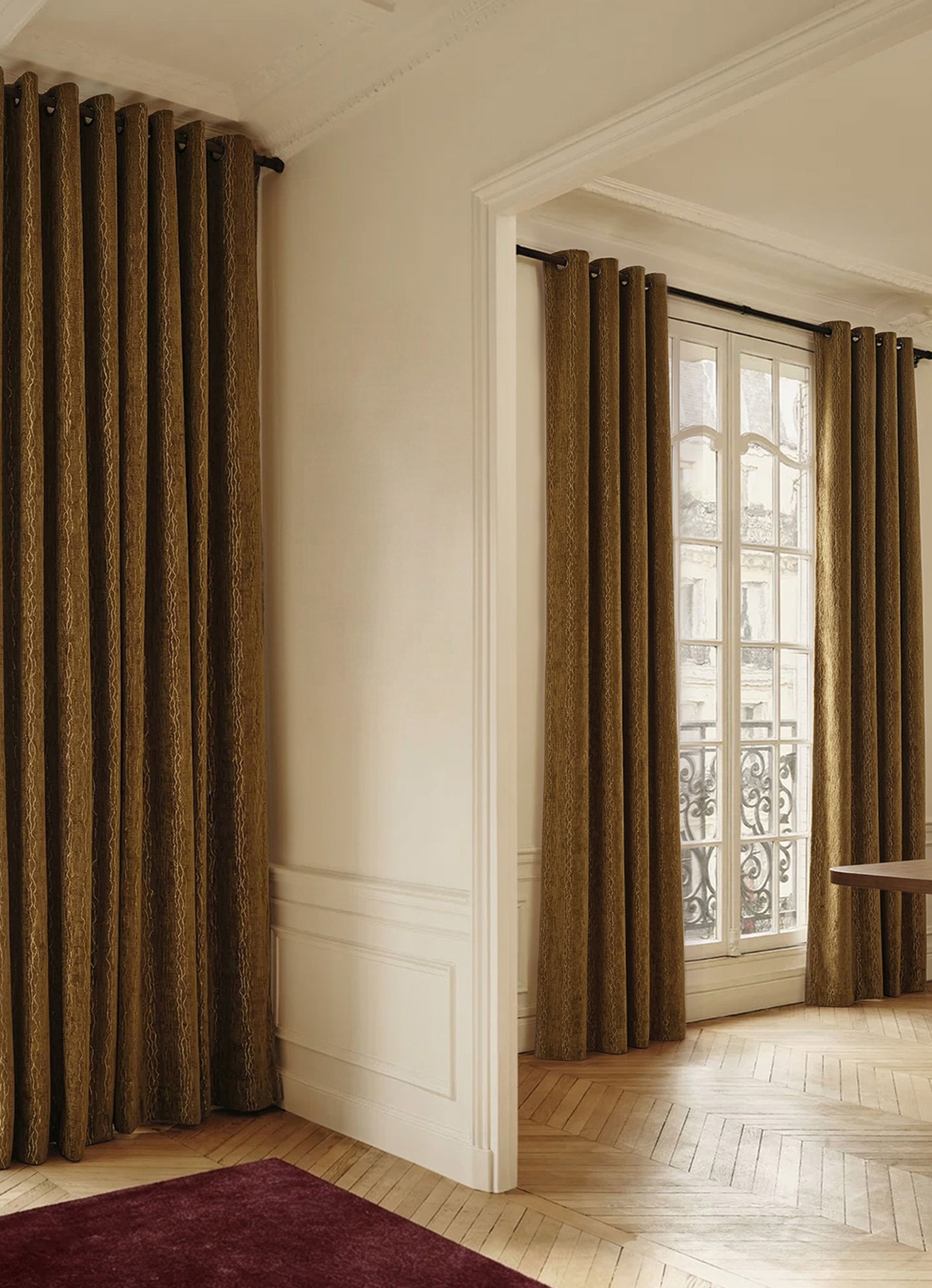 Room with large windows covered by brown patterned curtains, wood floors, and white walls, featuring a glimpse of an ornate balcony railing.