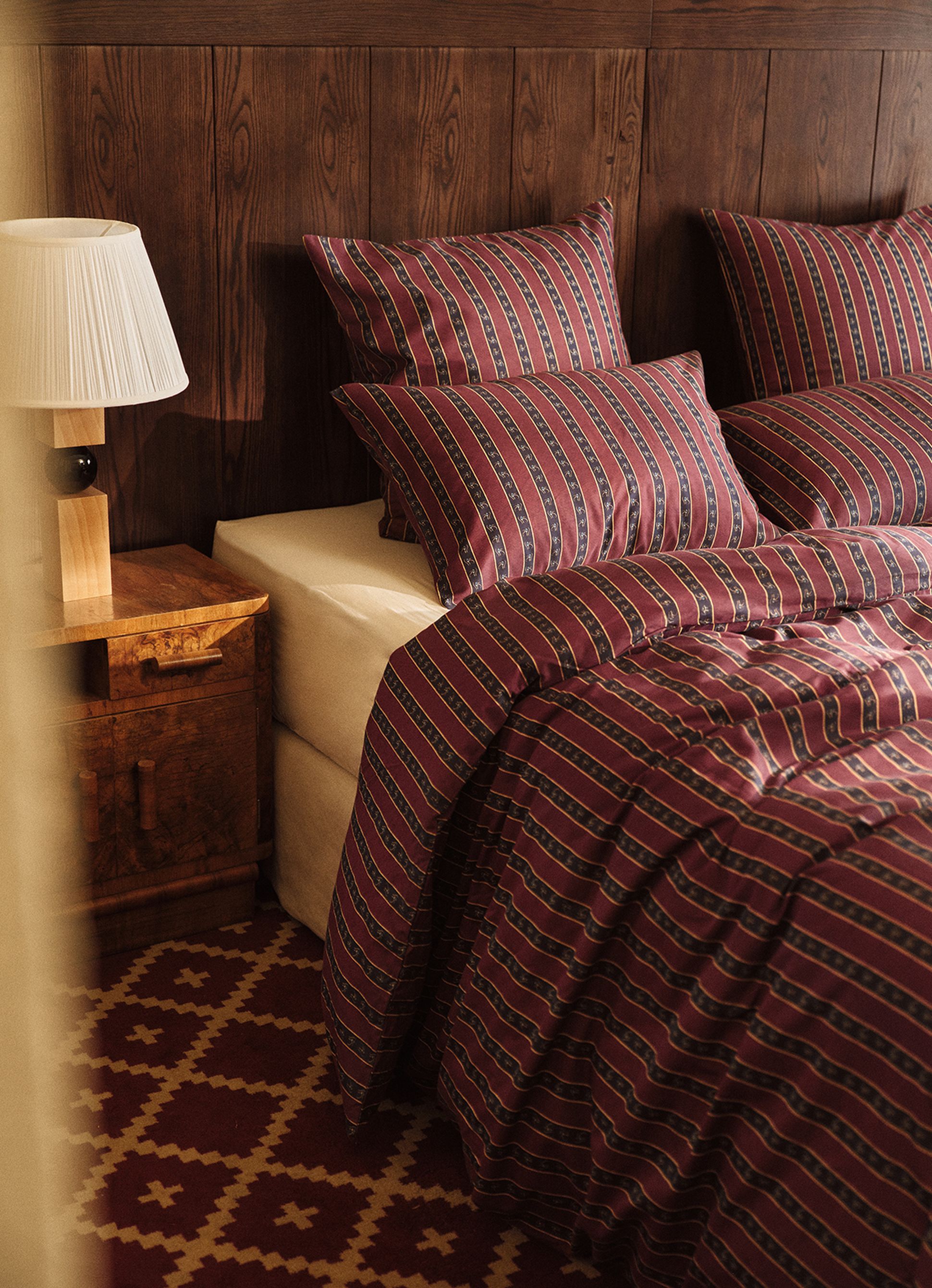 A cozy bedroom with a wooden wall, patterned burgundy bedding, and a bedside table with a lamp. The red and beige patterned rug adds warmth.
