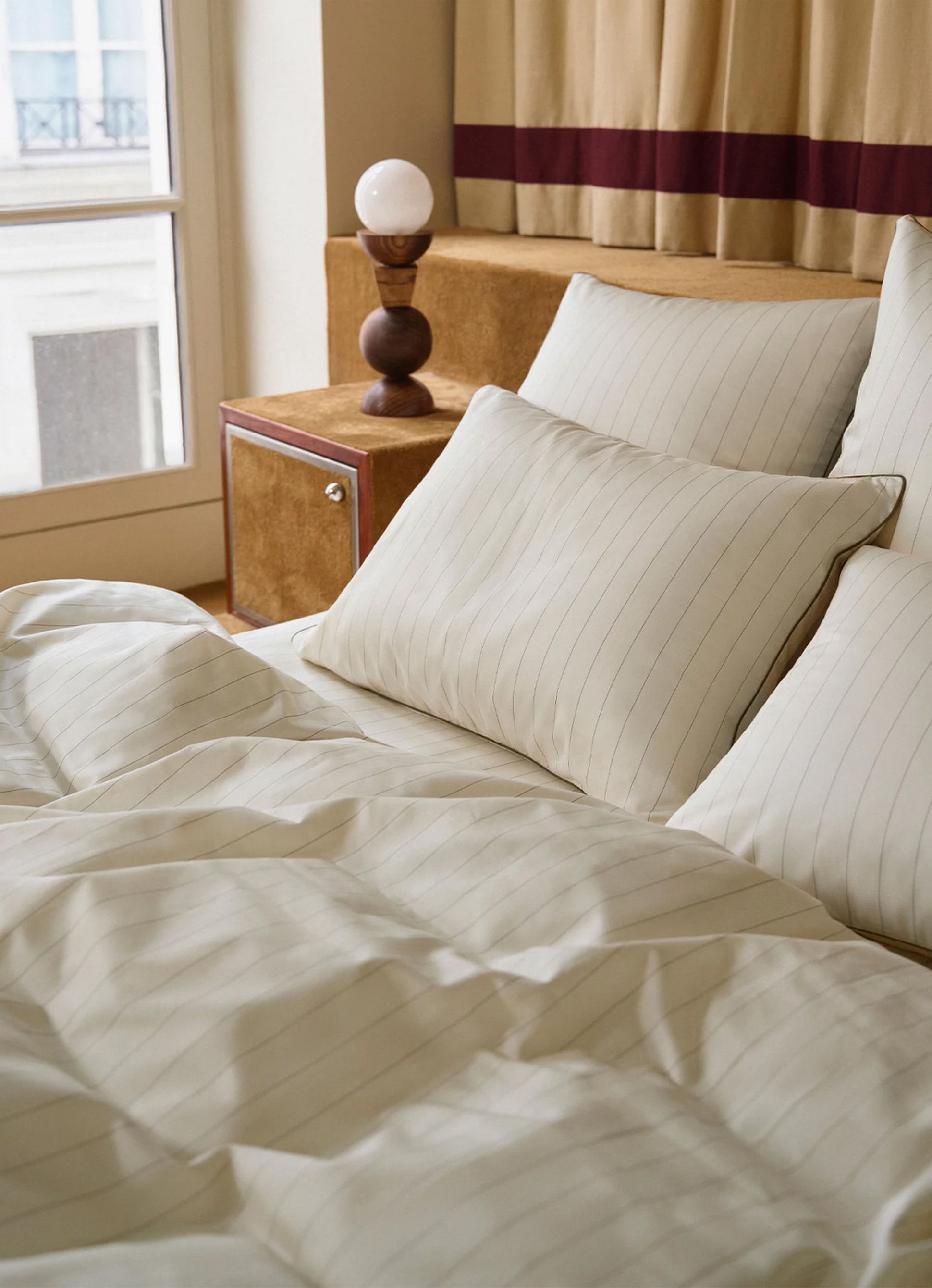 A cozy bed with striped white bedding and pillows, a wooden bedside table, a round lamp, and a window with light curtains.