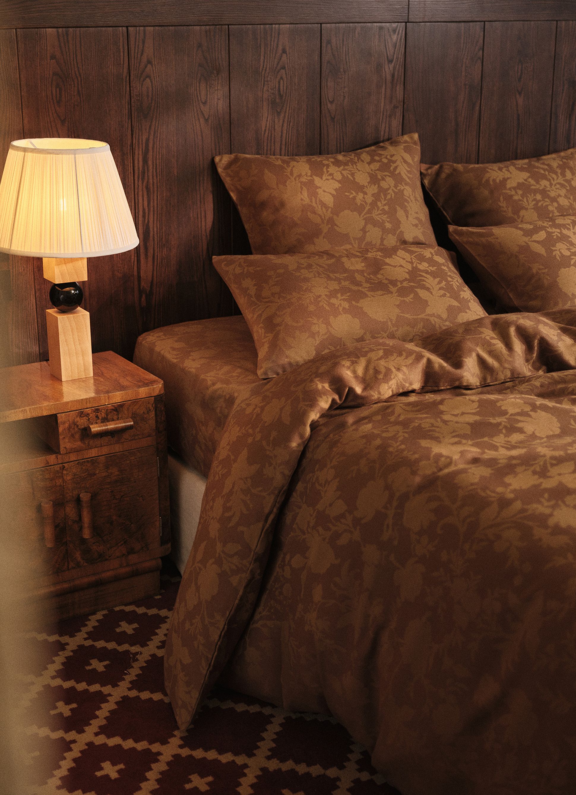 Une chambre douillette avec un mur lambrissé, un lit recouvert d'une parure de lit marron à motifs floraux, une table de chevet et une lampe allumée dotée d'un abat-jour plissé.