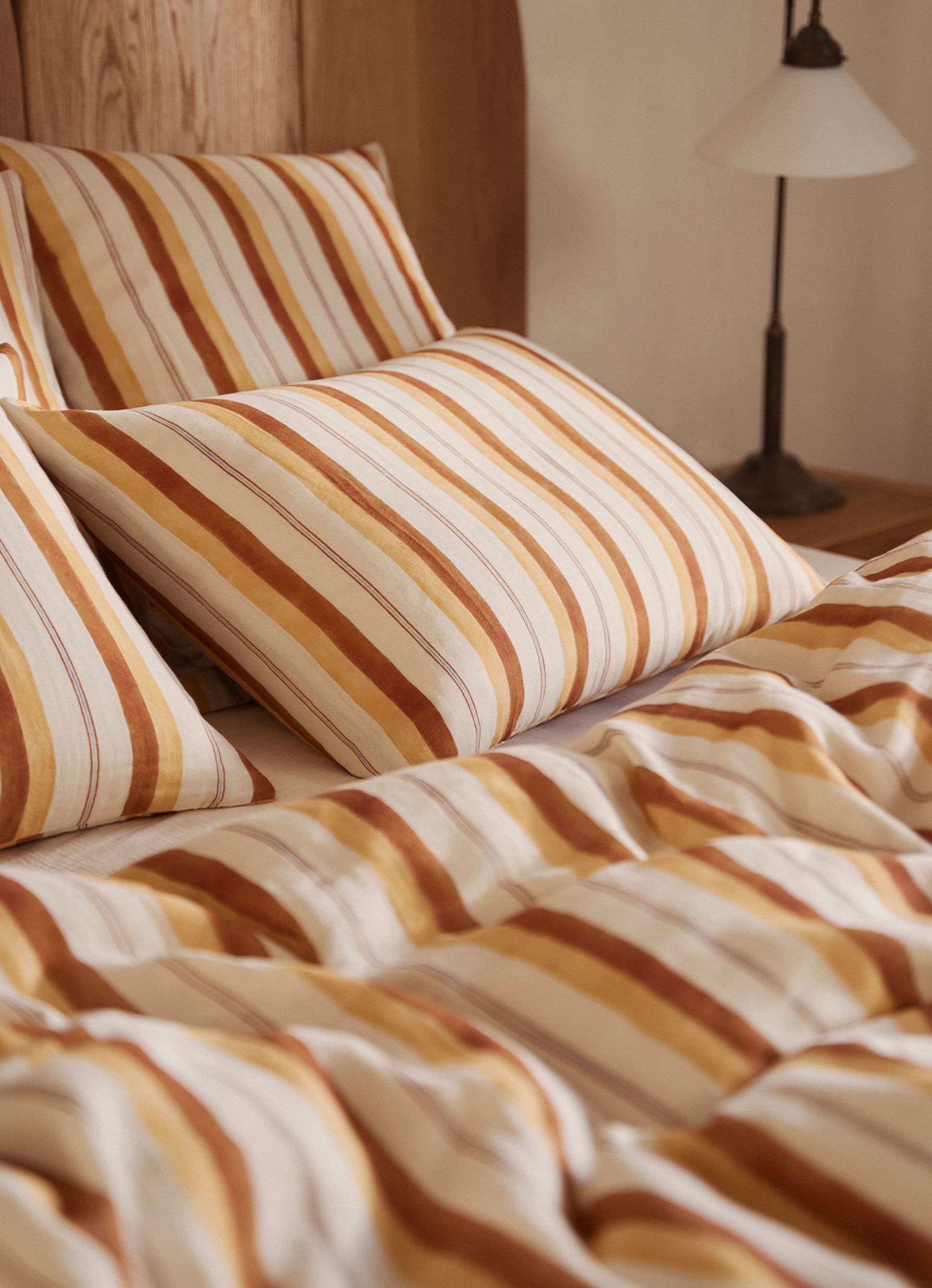 Un lit douillet avec des oreillers rayés et une couette dans les tons marron, orange et blanc. Une tête de lit en bois et une lampe se trouvent à l'arrière-plan.