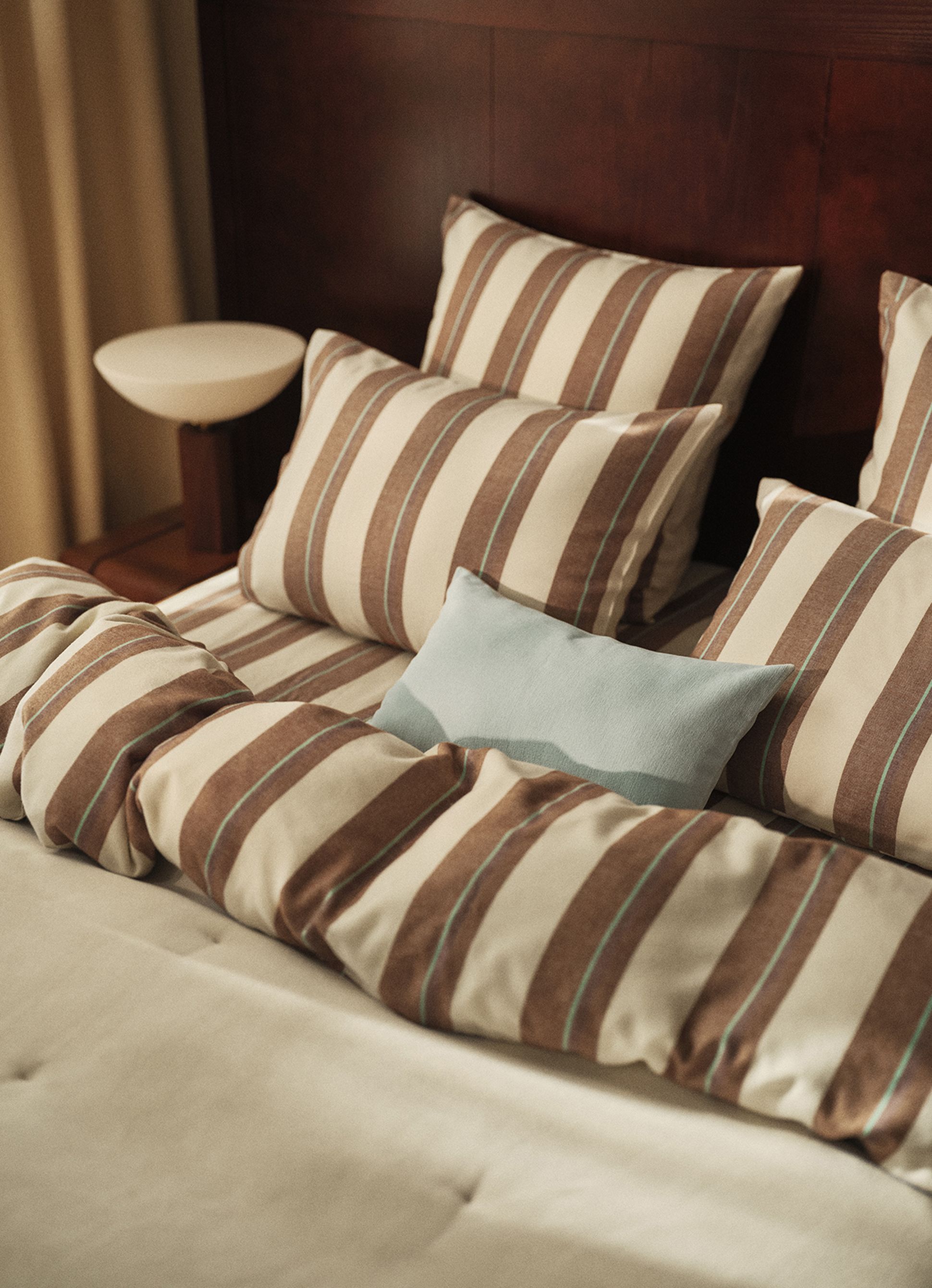 Un lit soigneusement fait, avec des oreillers et une couette rayés de marron et de beige, ainsi qu'un coussin décoratif bleu clair, le tout devant une tête de lit en bois foncé.