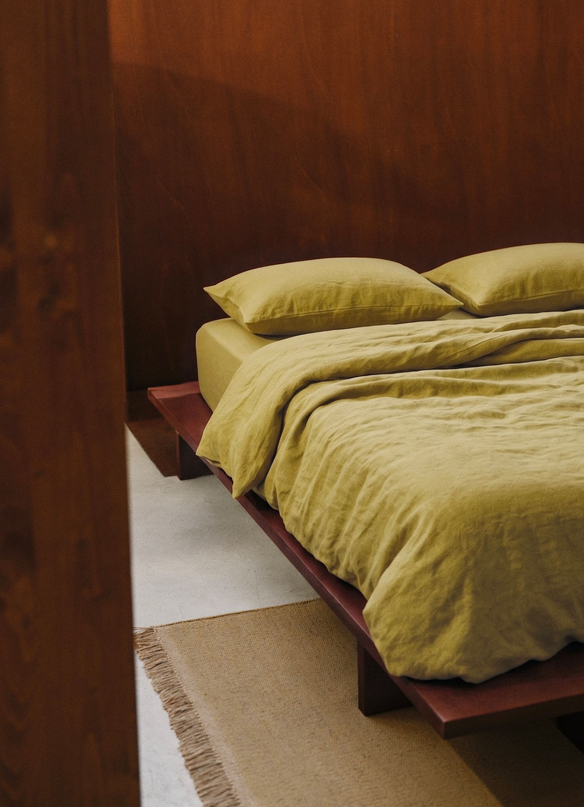 Une chambre minimaliste avec un lit surélevé en bois, recouvert d'une parure de lit jaune moutarde, posée sur un sol clair et texturé, agrémenté d'un tapis sobre.