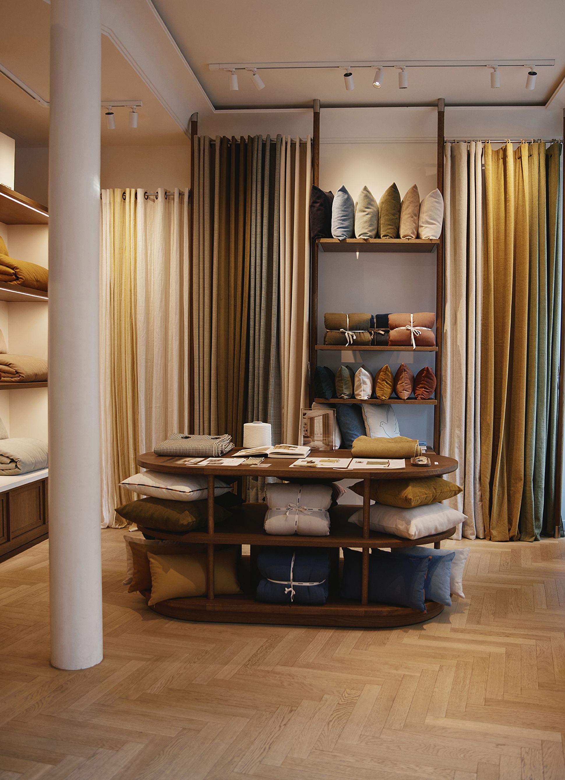 Une boutique de tissus chaleureuse, avec des étagères remplies de tissus colorés soigneusement pliés et de coussins, divers rideaux exposés, et un sol en bois à chevrons.