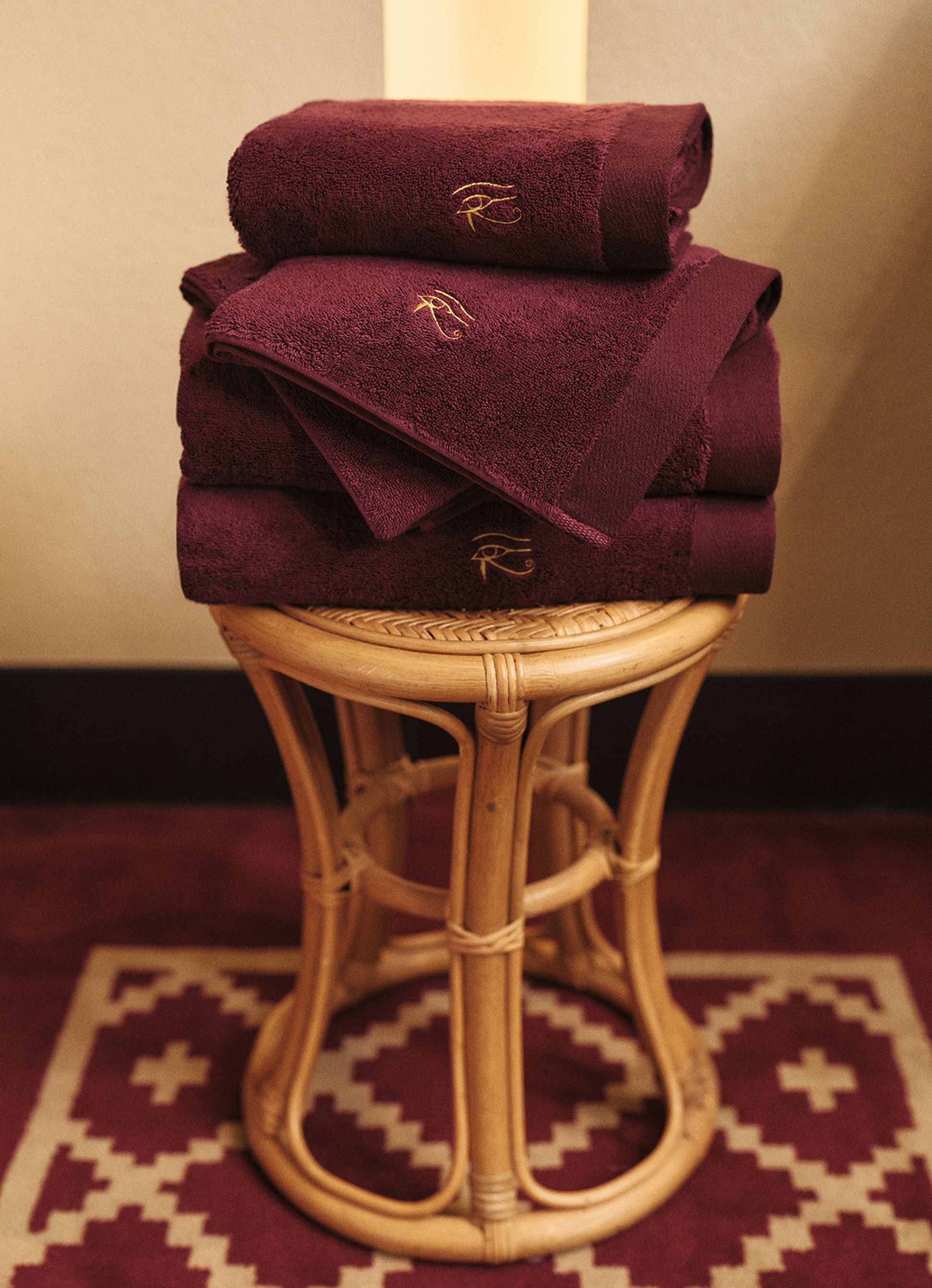 Stack of burgundy towels with embroidered logo on a wicker stool, set against a patterned rug.
