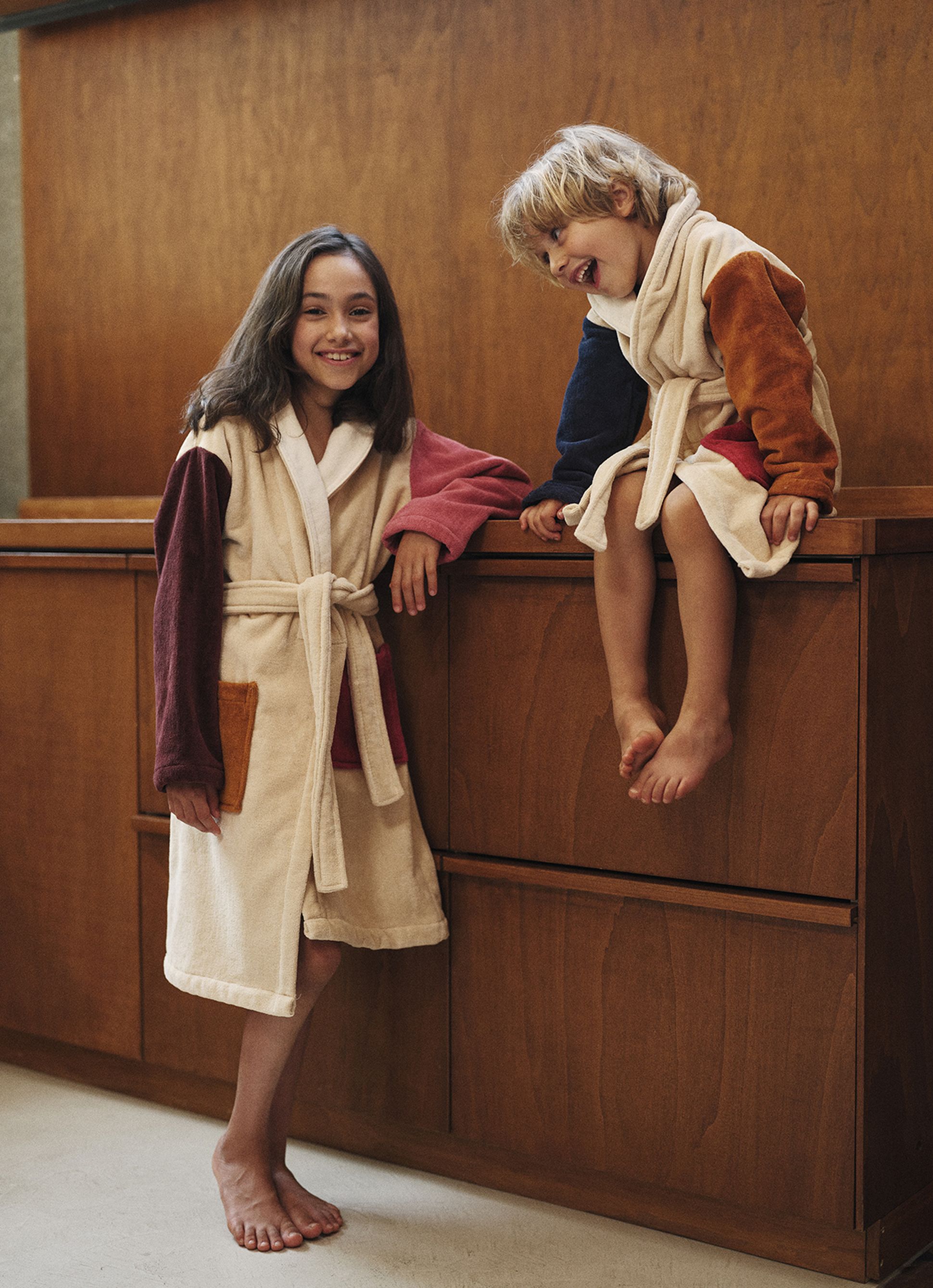 Deux enfants vêtus de robes colorées, l'un debout et l'autre assis sur un meuble en bois, sourient chaleureusement dans un intérieur chaleureux.