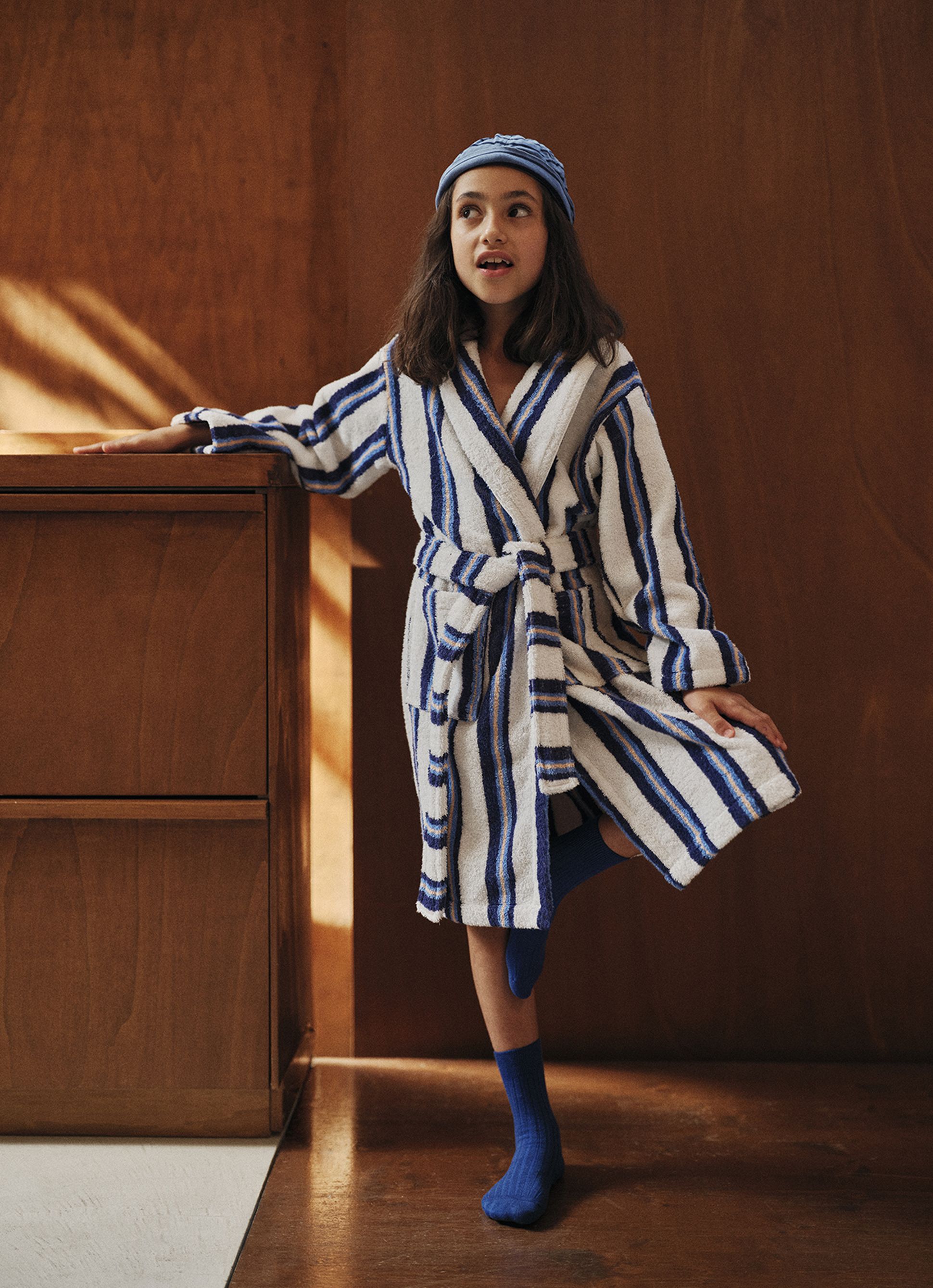 Une jeune fille vêtue d'une robe rayée bleue et blanche et d'un bonnet bleu se tient à l'intérieur, adossée à une commode en bois, l'air intrigué.