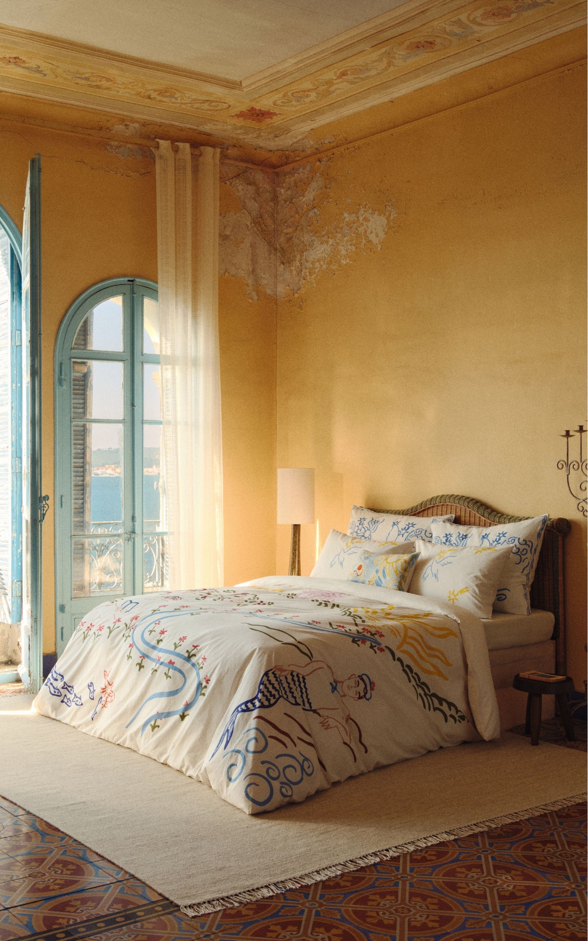 Une chambre baignée de soleil, avec une parure de lit blanche ornée de motifs abstraits colorés, une tête de lit en osier et un grand lampadaire placé près d'une fenêtre aux rideaux vaporeux.