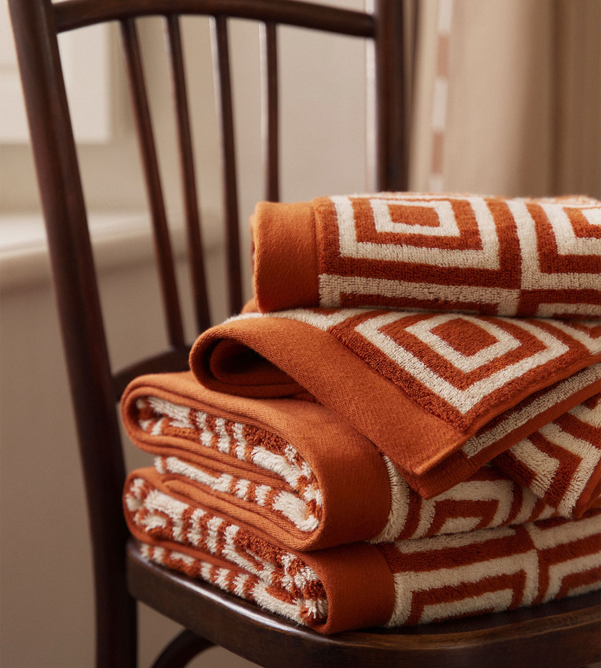 Pile de serviettes orange à motifs géométriques blancs sur une chaise en bois, placée à côté d'une fenêtre à rideaux dans une lumière tamisée.