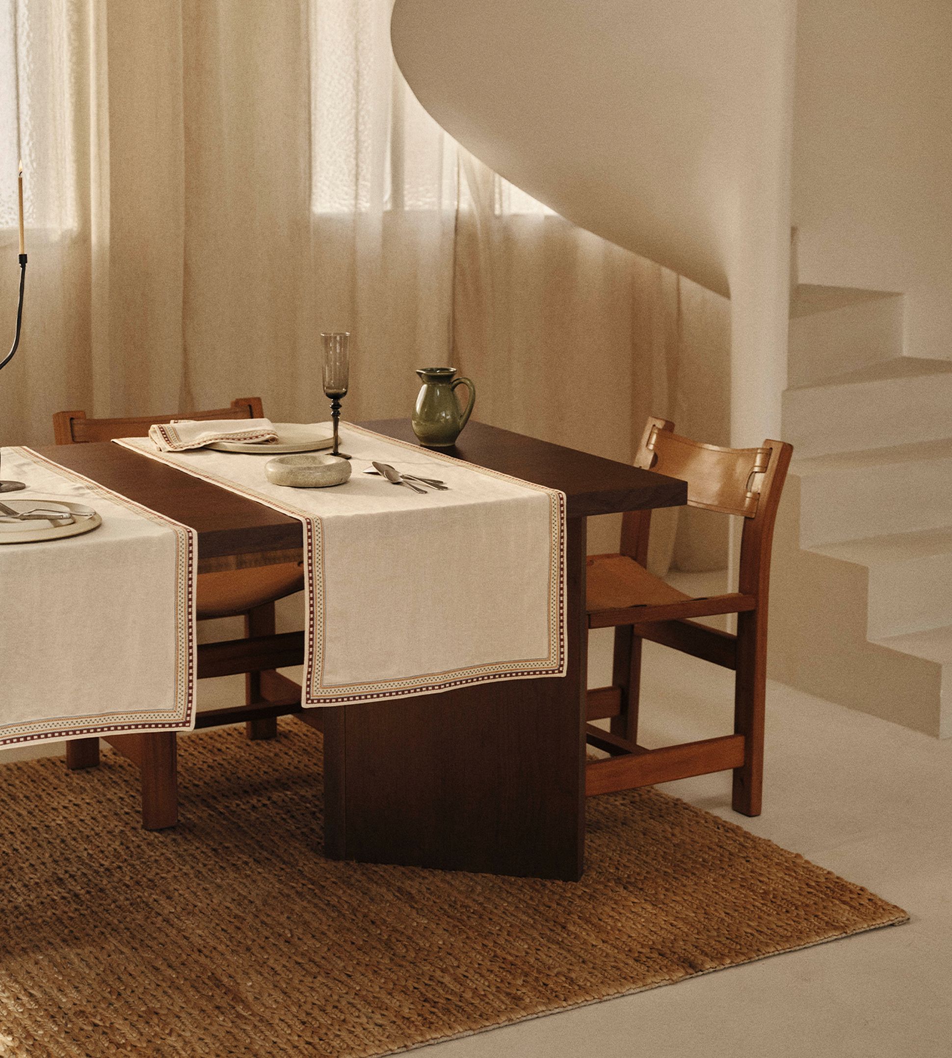 Une table à manger en bois avec des sets de table blancs, un verre et un pichet. Elle se trouve dans une pièce baignée de lumière naturelle, avec un escalier en colimaçon en arrière-plan.