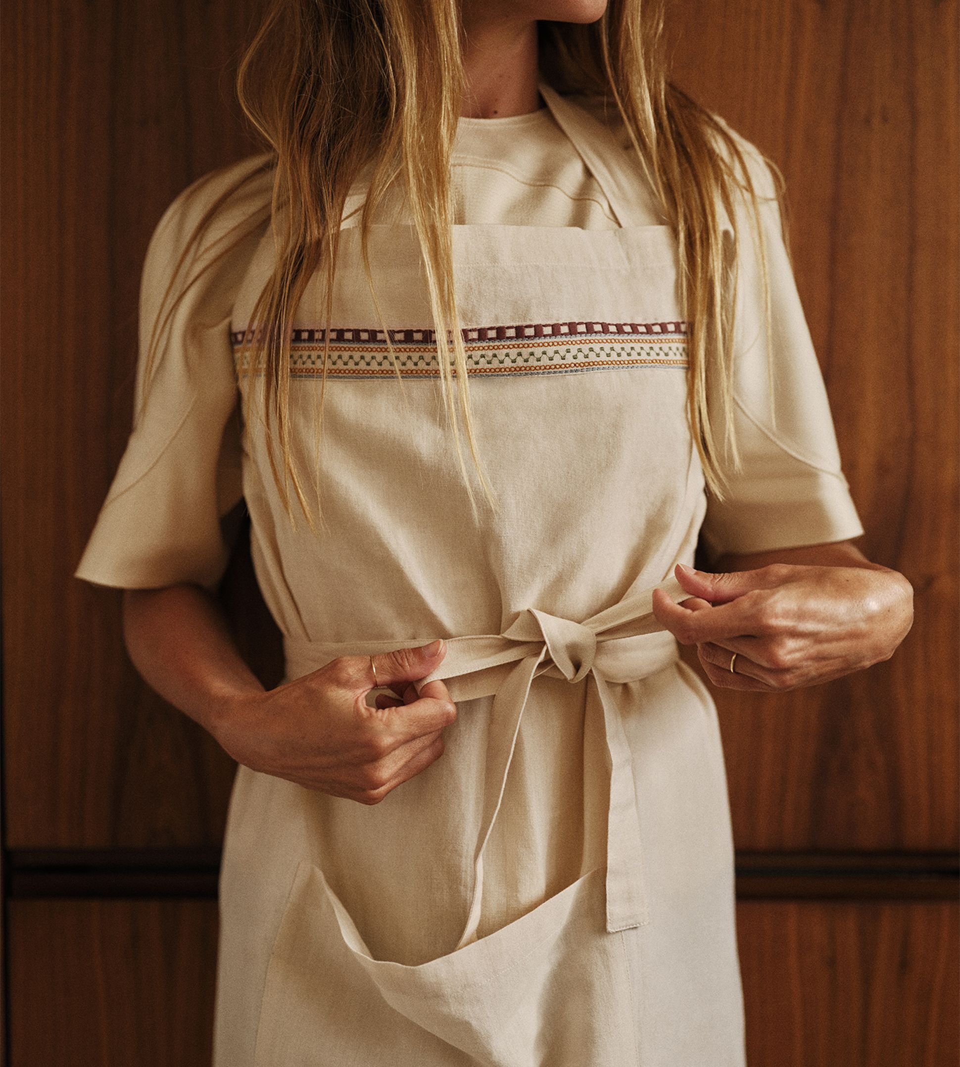 Une femme en train de faire un nœud sur un tablier beige orné de surpiqûres décoratives, debout devant un fond en bois.