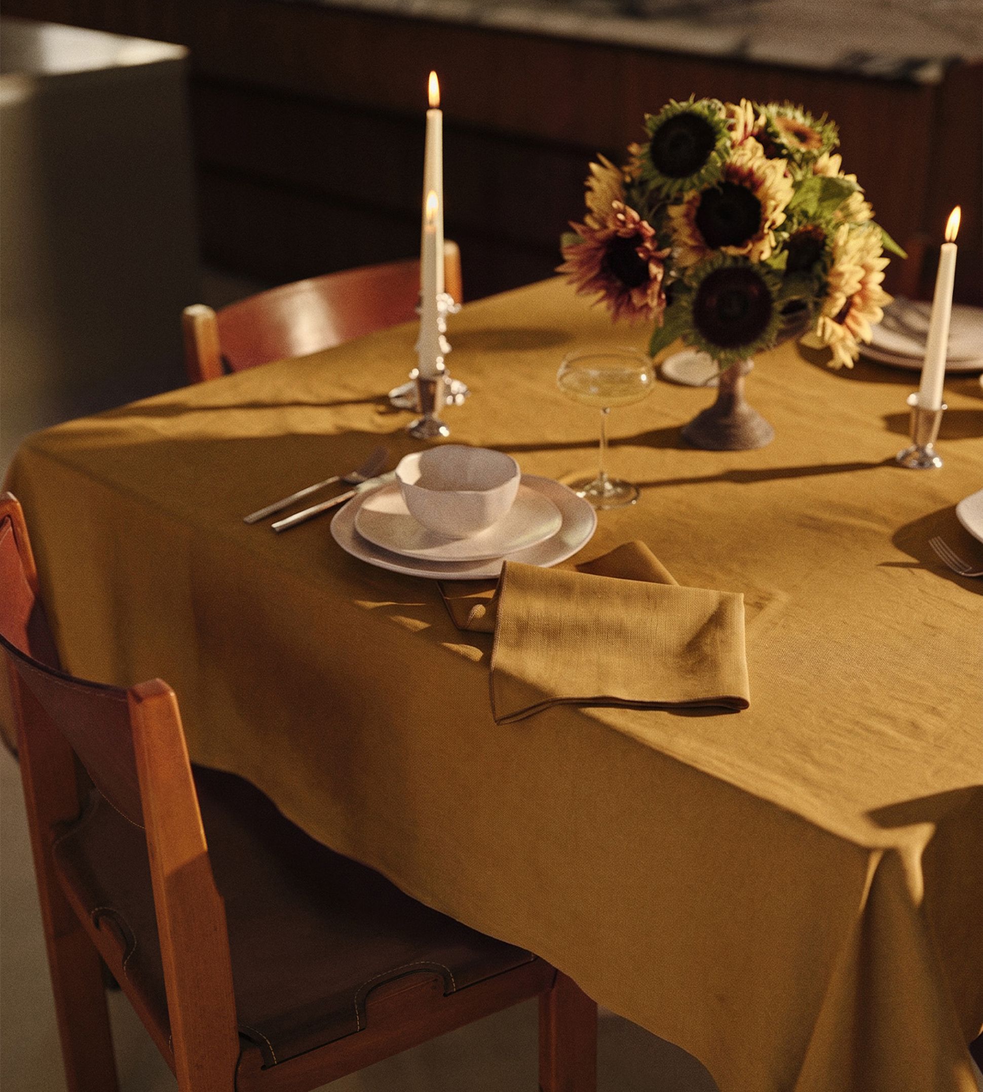 Une table à manger baignée d'une lumière chaleureuse, recouverte d'une nappe couleur moutarde, dressée avec un bol et une assiette, des couverts, une bougie allumée, une serviette pliée et un vase rempli de tournesols.