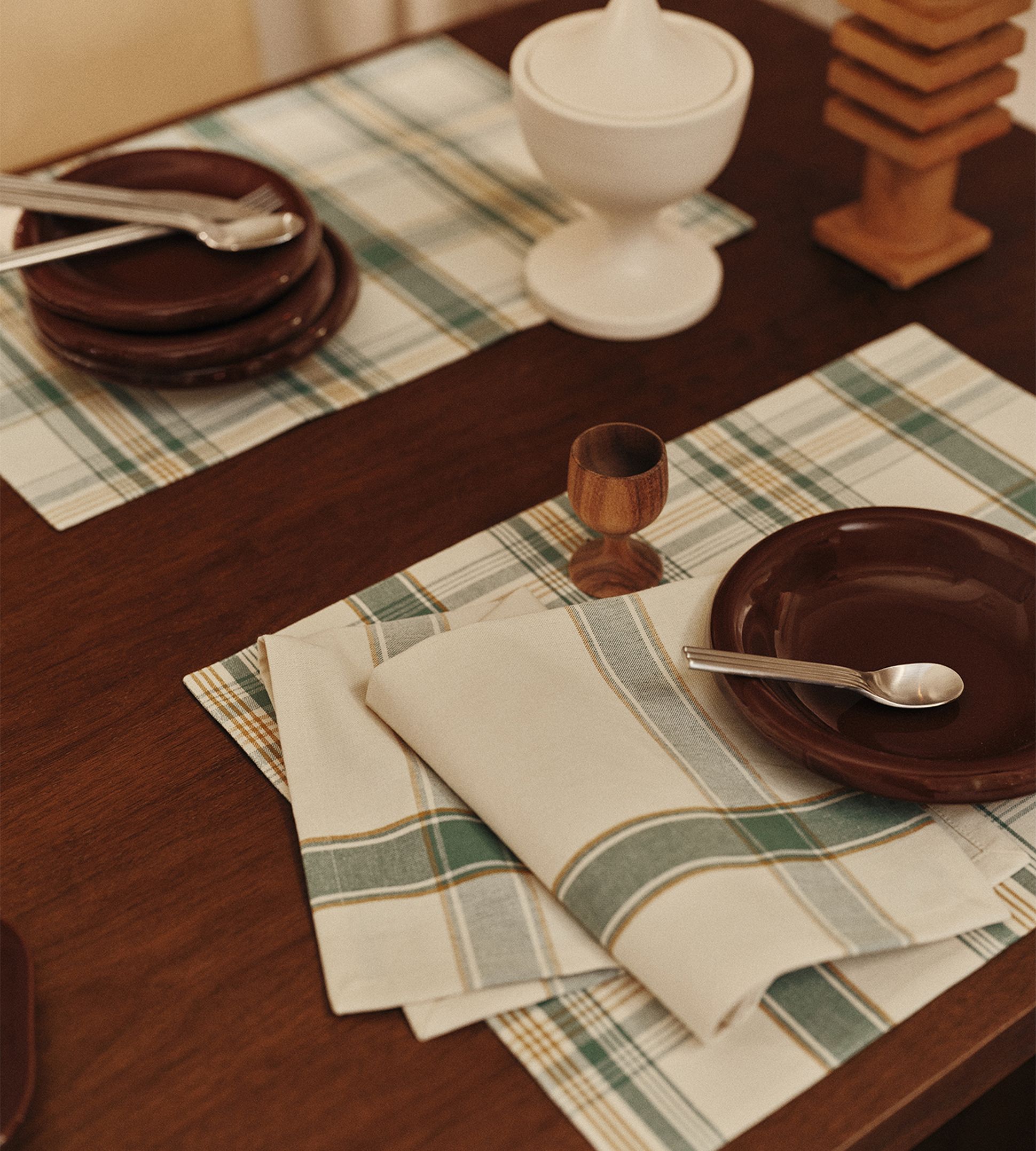 Une table dressée avec un set de table à carreaux, une assiette marron, une cuillère, une serviette pliée, un coquetier en bois et un bol muni d'un couvercle, dans une salle à manger aux tons chauds.