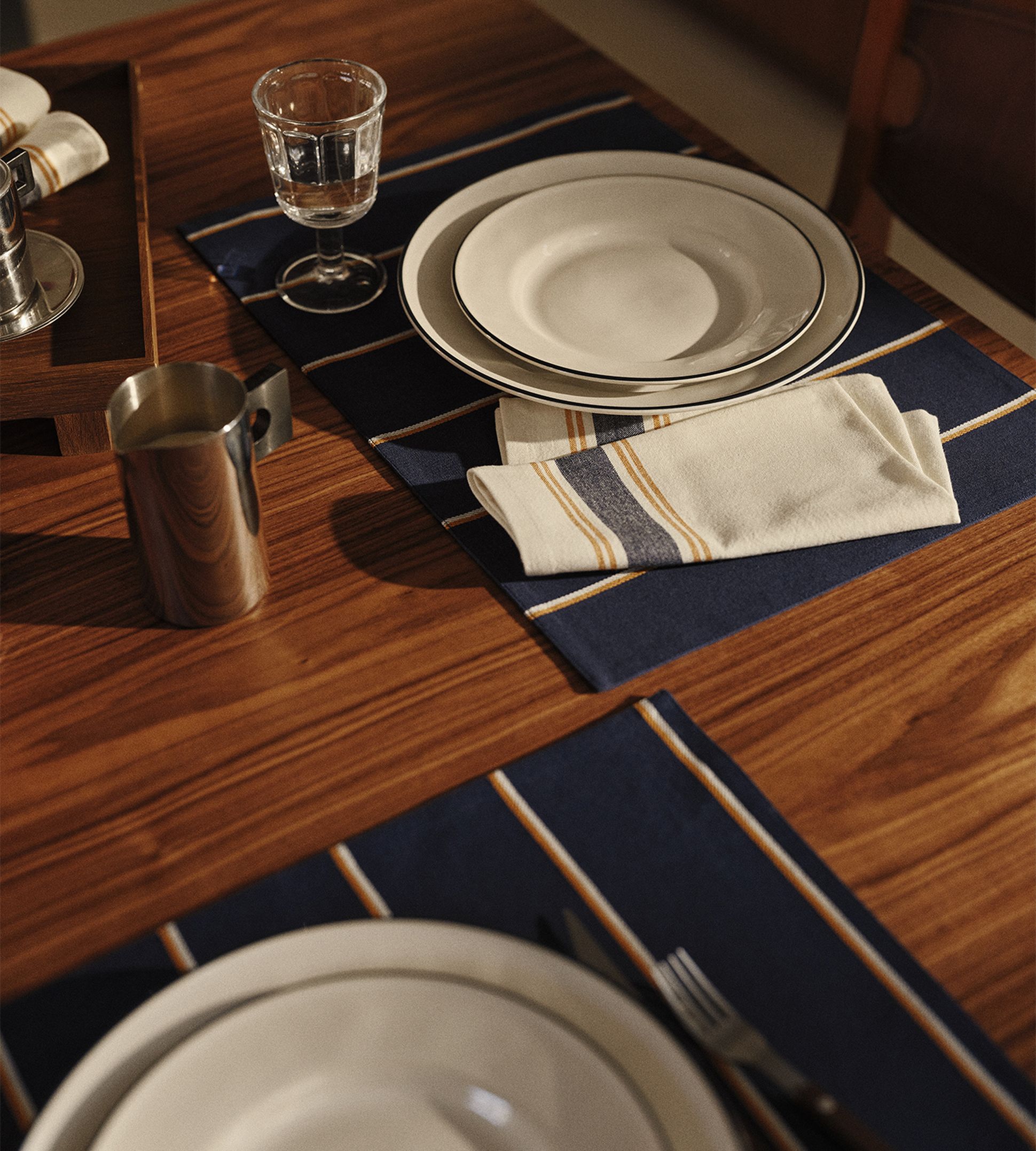 Une table élégamment dressée avec des assiettes blanches posées sur des sets de table foncés, une serviette rayée pliée, un verre à eau et une tasse en métal sur une table en bois.