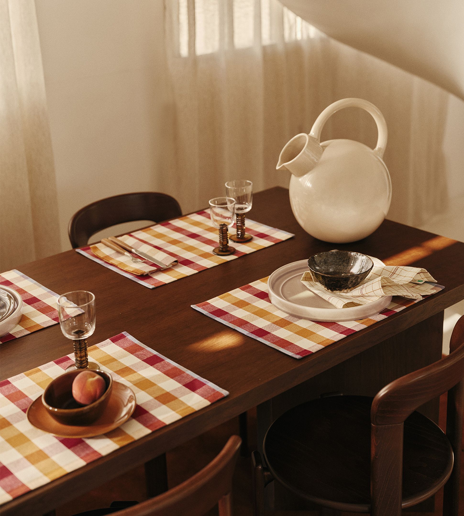 Une table à manger dressée avec des sets de table à carreaux, des verres, un pichet rond blanc et des bols, baignée d'une douce lumière naturelle filtrant à travers des rideaux vaporeux.