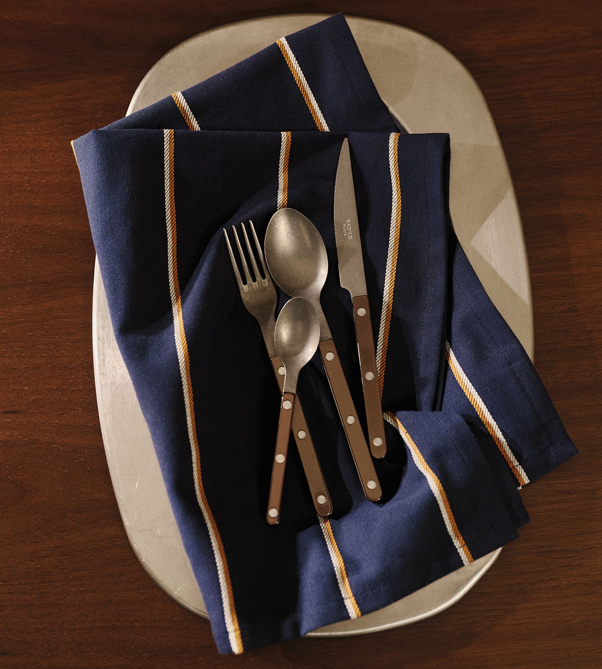 Un service de couverts aux manches marron posé sur une serviette à rayures bleues, lui-même posé sur une assiette, le tout disposé sur une table en bois.
