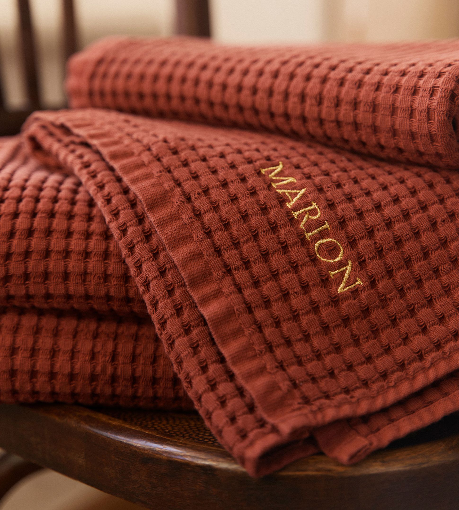 Pile de serviettes en maille gaufrée couleur rouille avec « MARION » brodé en doré, posée sur une chaise en bois.
