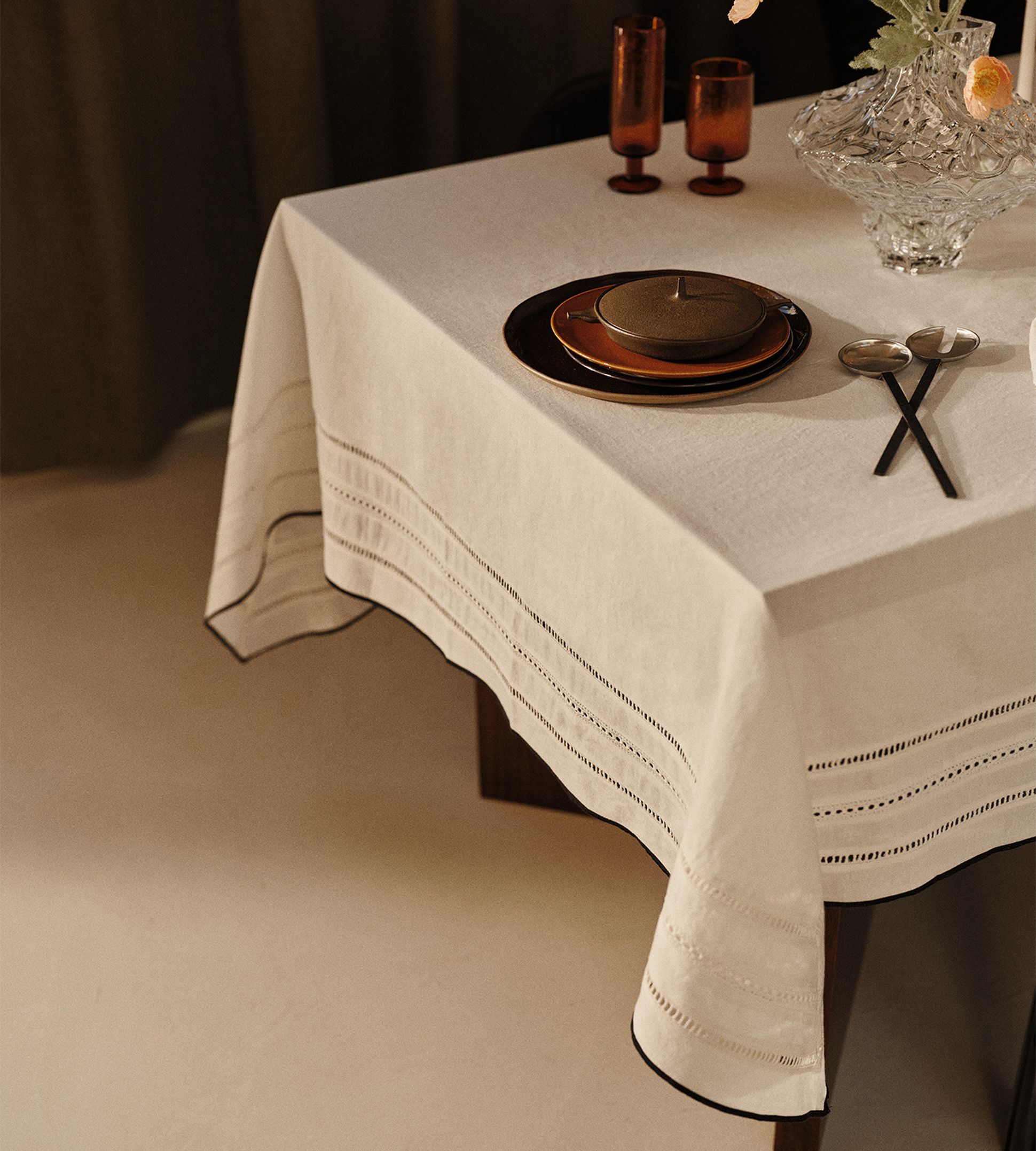 Une table élégamment dressée, avec une nappe blanche, de la vaisselle marron, un vase en cristal orné de fleurs et deux cuillères posées sur la table.