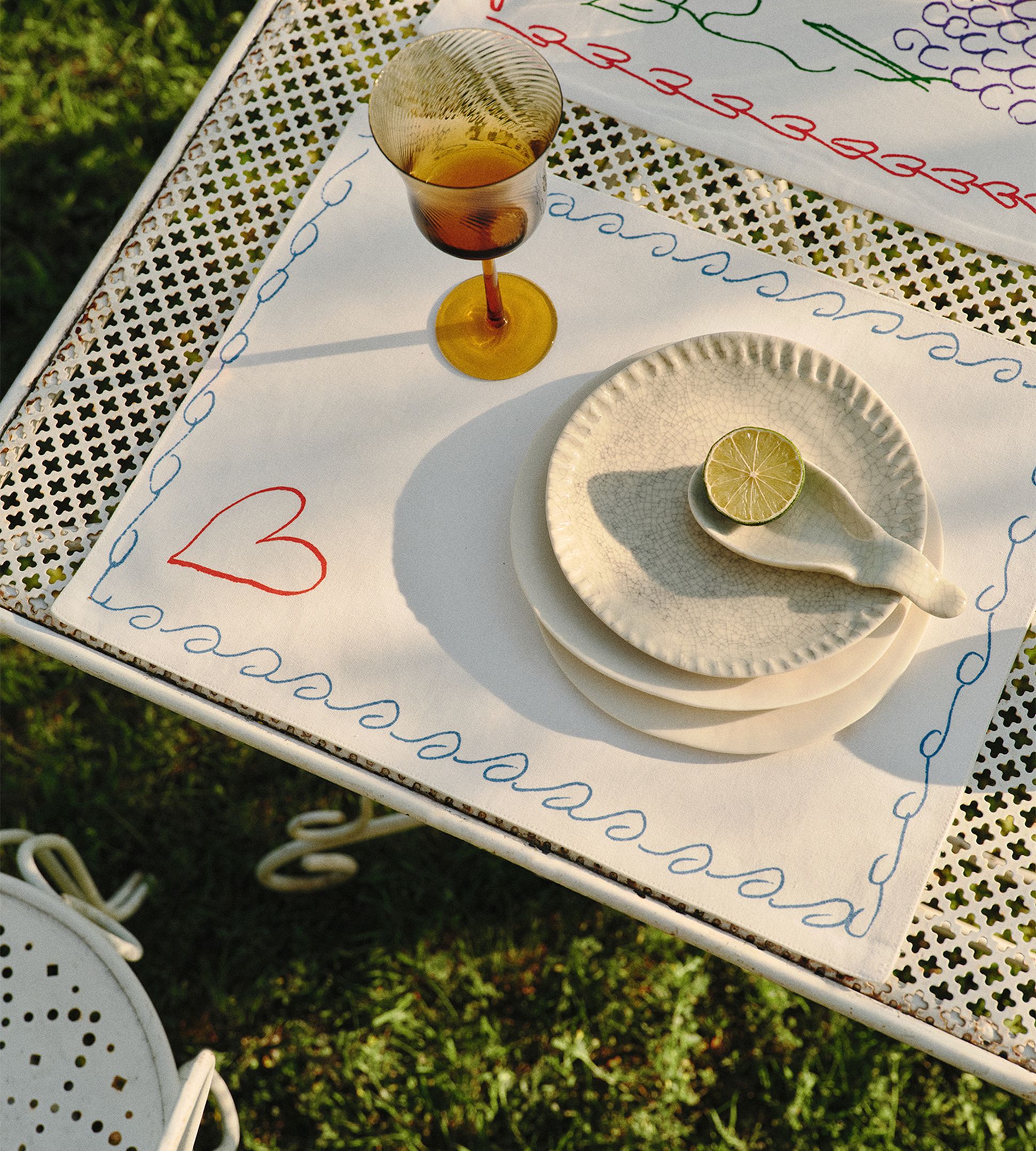 Une table en métal dressée à l'extérieur, avec des sets de table à motifs floraux, un calice jaune et des assiettes en céramique empilées, surmontées de tranches de citron vert.