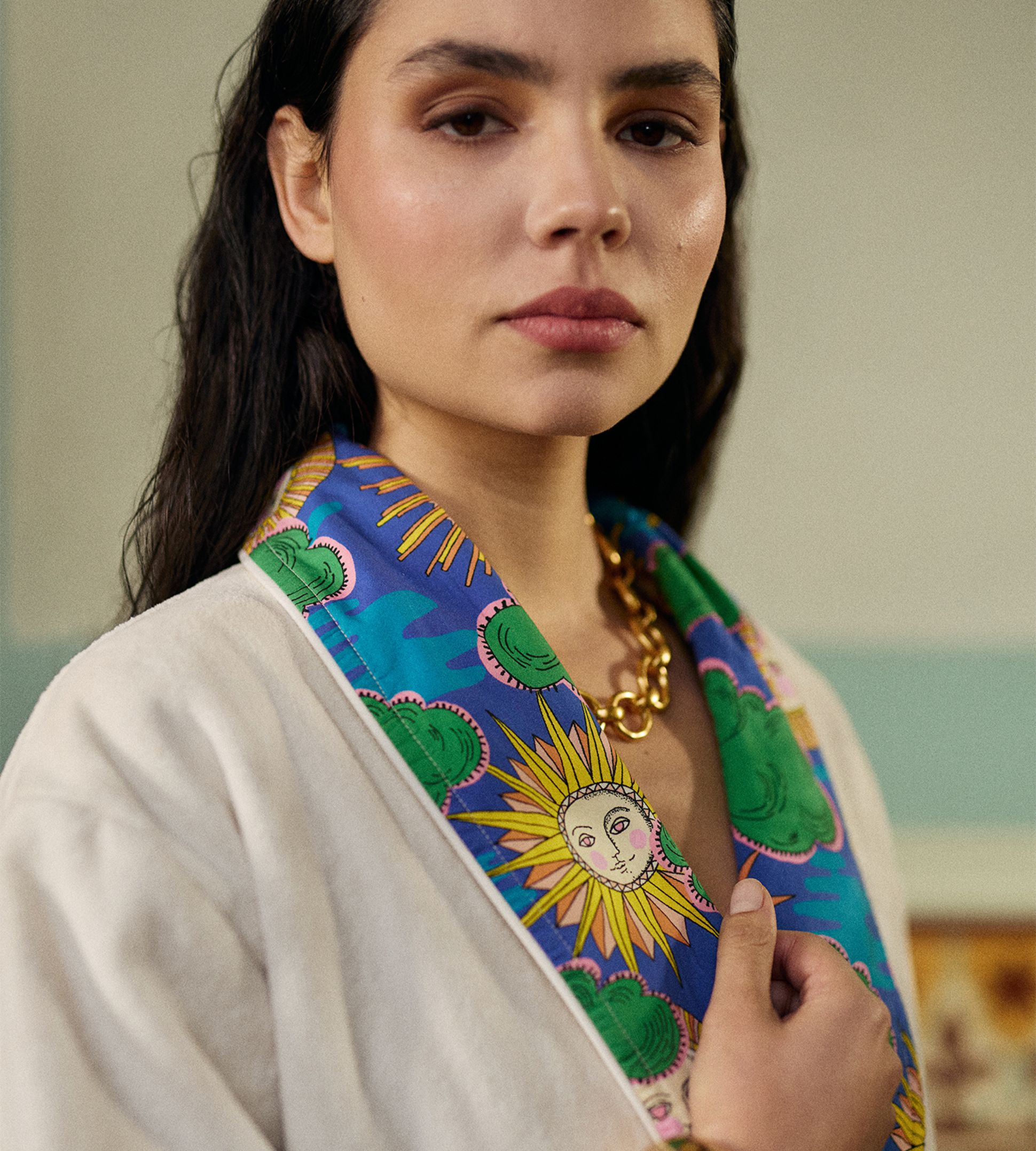 Une femme vêtue d'un peignoir blanc orné d'un col coloré à motifs de soleil, portant un gros bracelet et un collier en or, le regard assur