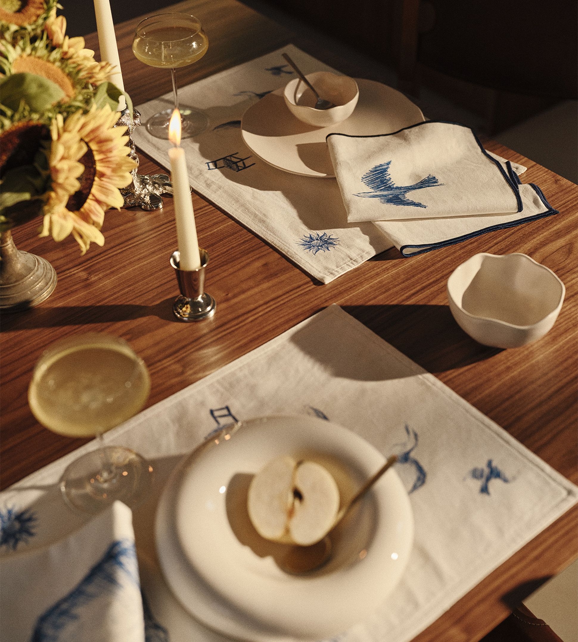 Une table en bois dressée avec des assiettes blanches, une pomme coupée en deux, des coupes à champagne, des serviettes brodées, des tournesols et une bougie allumée.