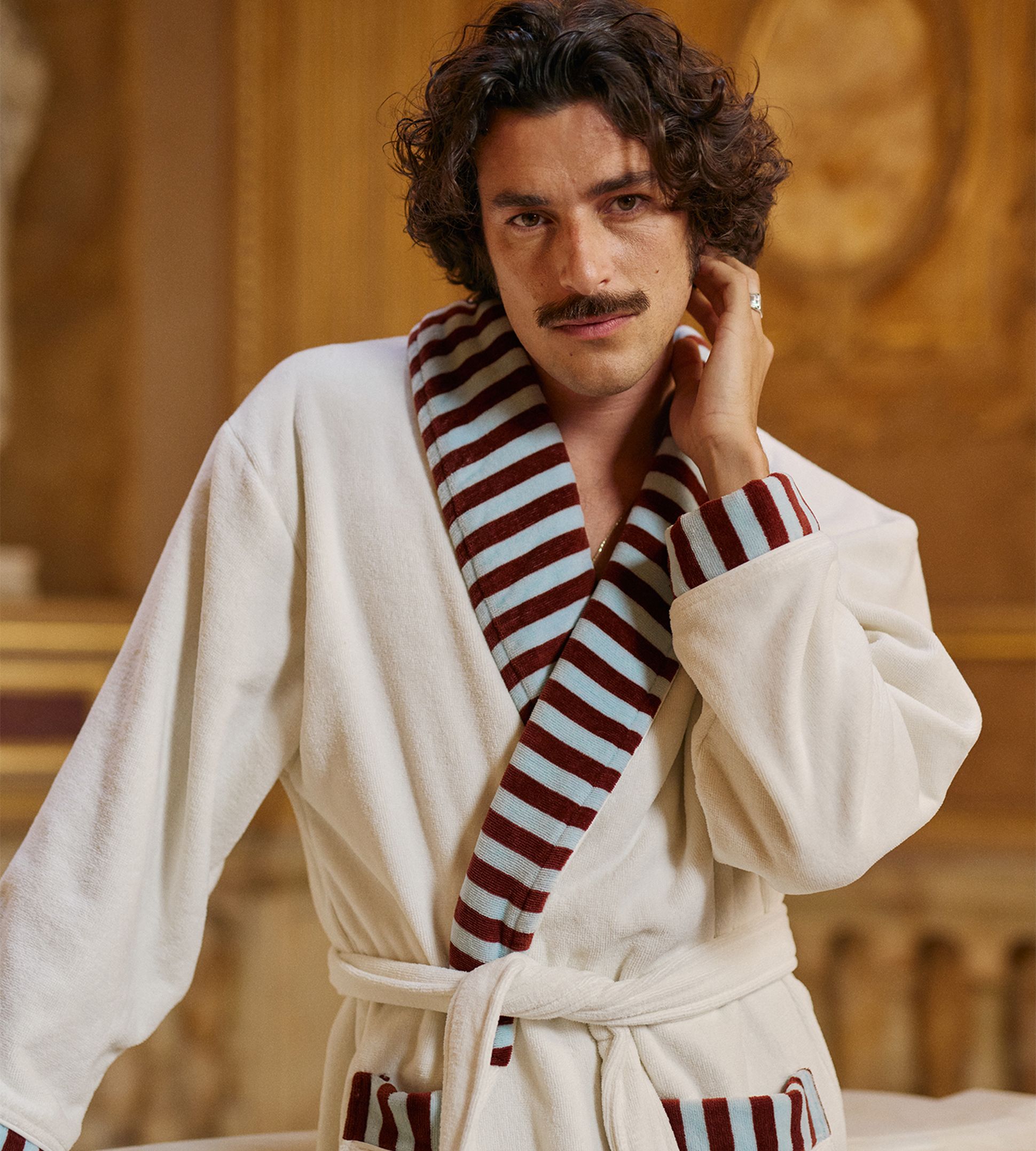 Un homme aux cheveux bouclés et à la moustache, vêtu d'un peignoir blanc à rayures rouges et bleues, posant dans un décor raffiné.