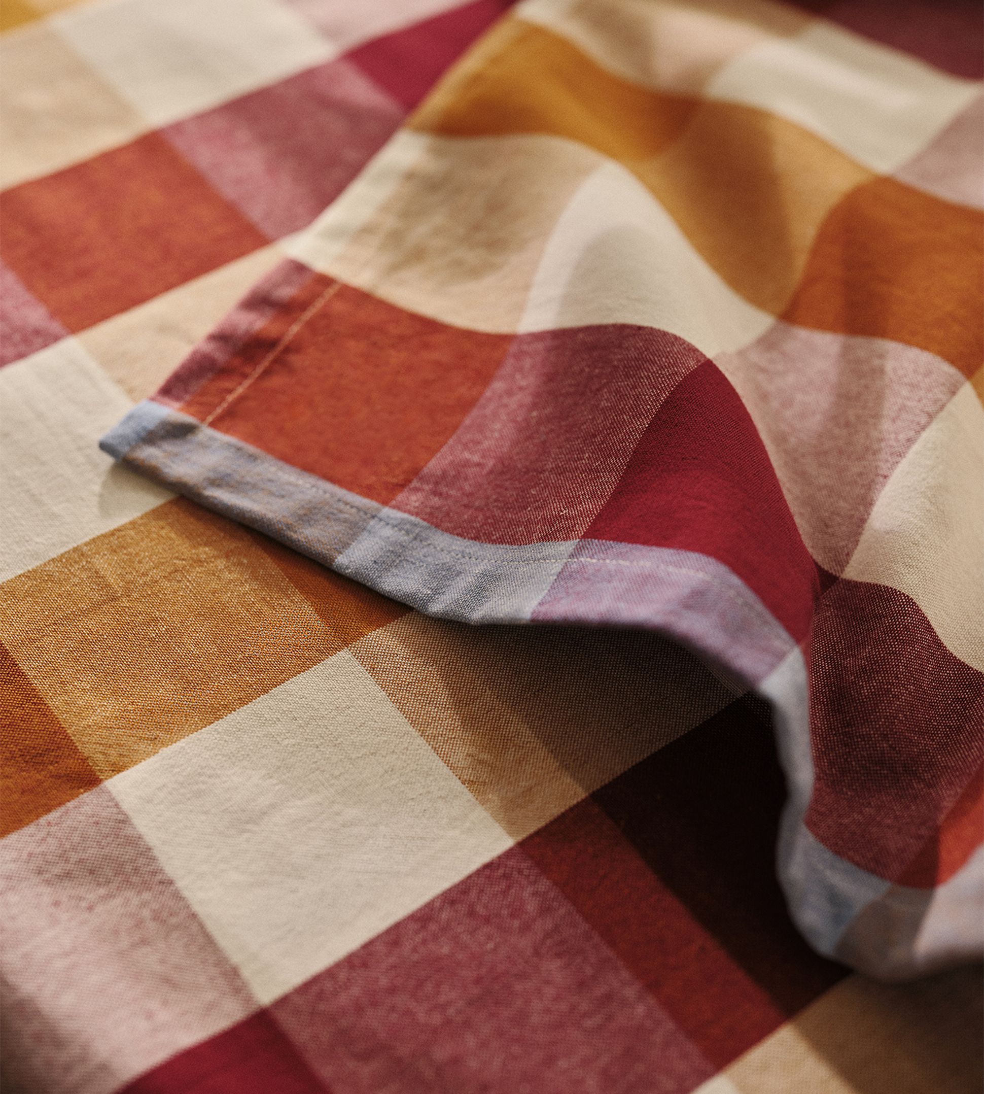 Gros plan sur un tissu plié présentant un grand motif à carreaux rouges, orange et beiges, bordé de bleu clair.