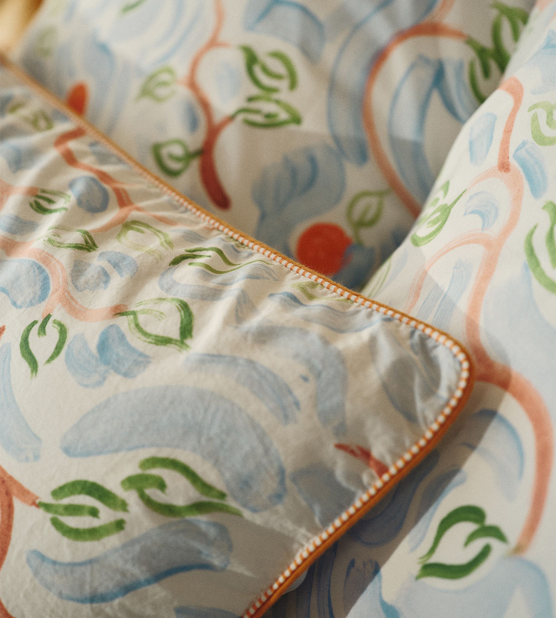 Gros plan sur des coussins au motif abstrait composé de lignes courbes bleues, vertes et orange, rehaussés d'un liseré orange.