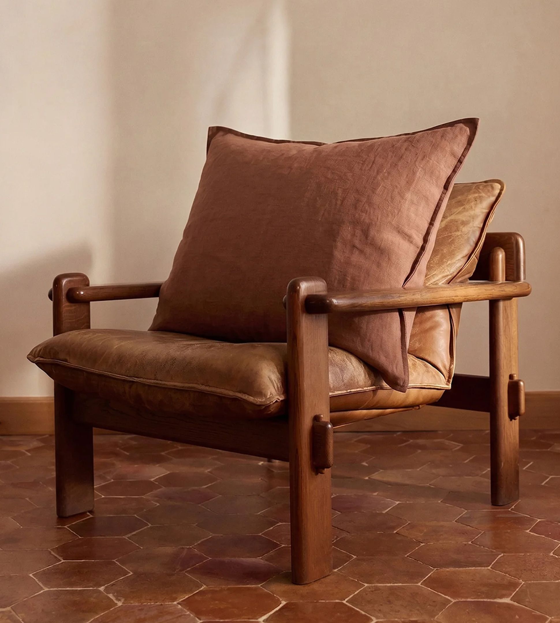 Une chaise en bois avec des coussins en cuir marron et un grand coussin marron, posée sur un sol en carrelage hexagonal, adossée à un mur beige.