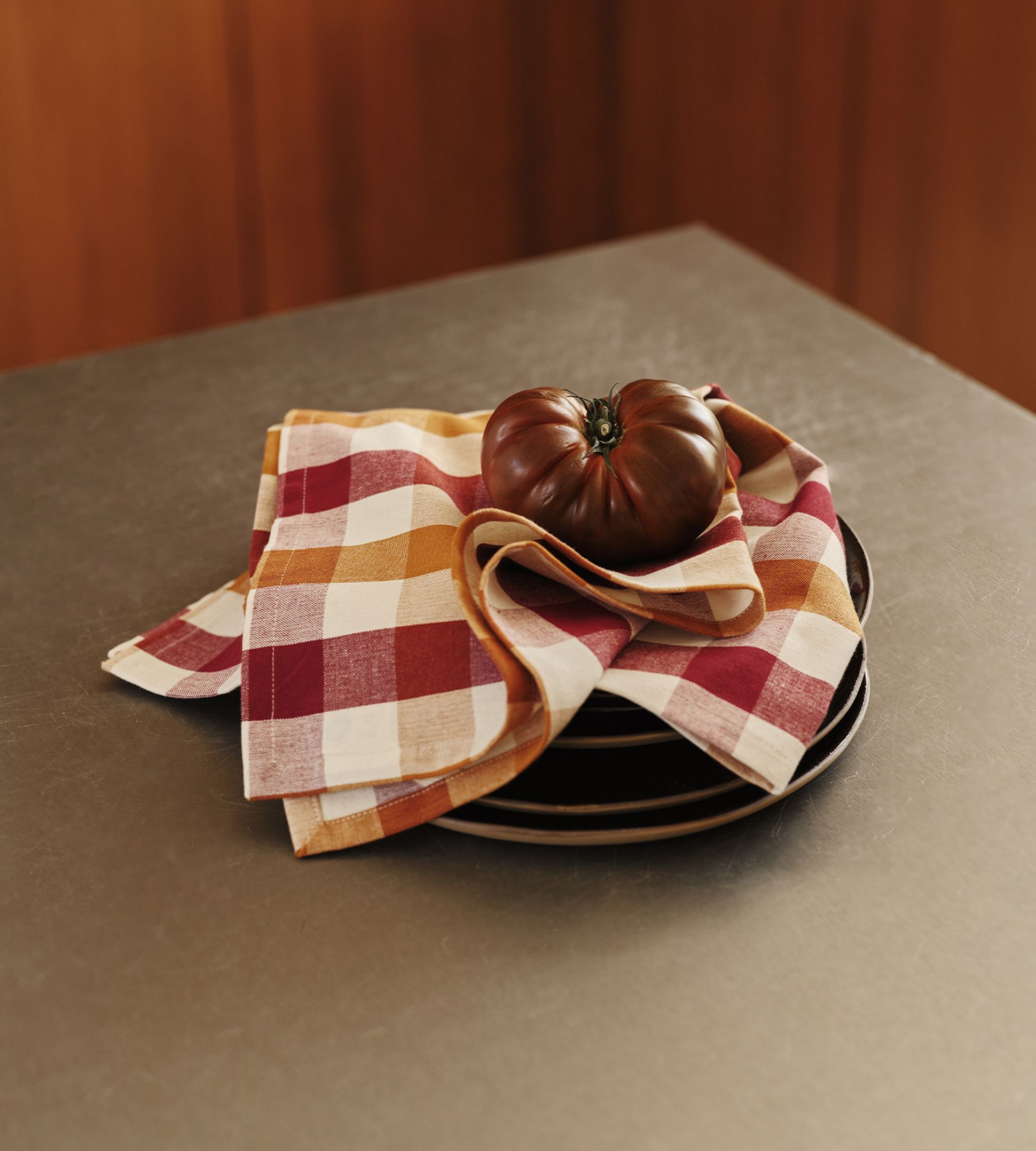 Une tomate foncée repose sur une pile d'assiettes recouverte d'une serviette à carreaux rouges, orange et blancs, posée sur un plan de travail.