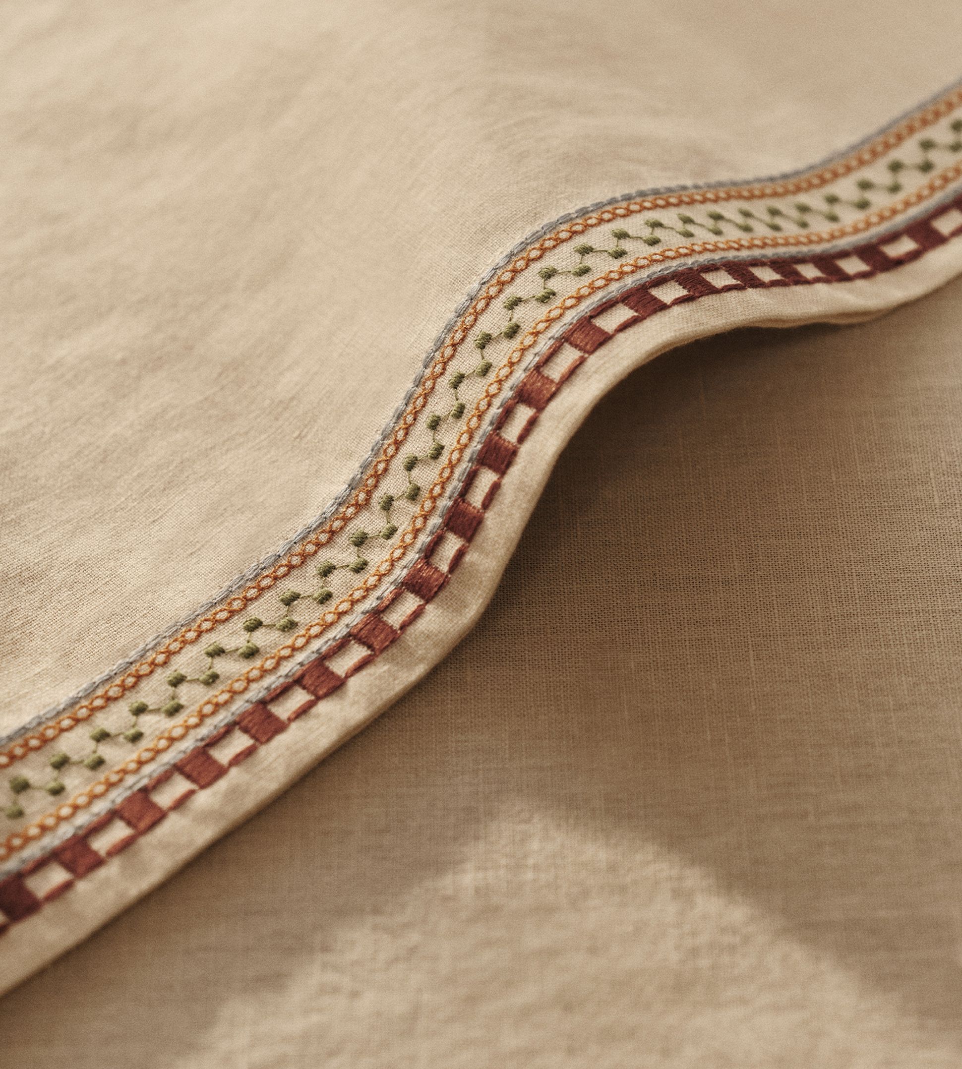 Gros plan sur un tissu beige orné de broderies détaillées en rouge, vert et orange, mettant en valeur des motifs complexes le long du bord.
