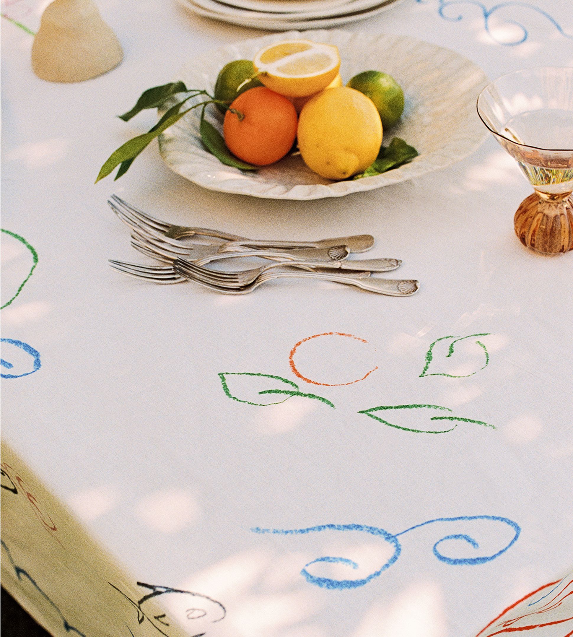 Une table recouverte d'une nappe blanche ornée de motifs de feuilles colorées, dressée avec des fourchettes et une corbeille de fruits contenant des agrumes, qui projettent de douces ombres tachetées.