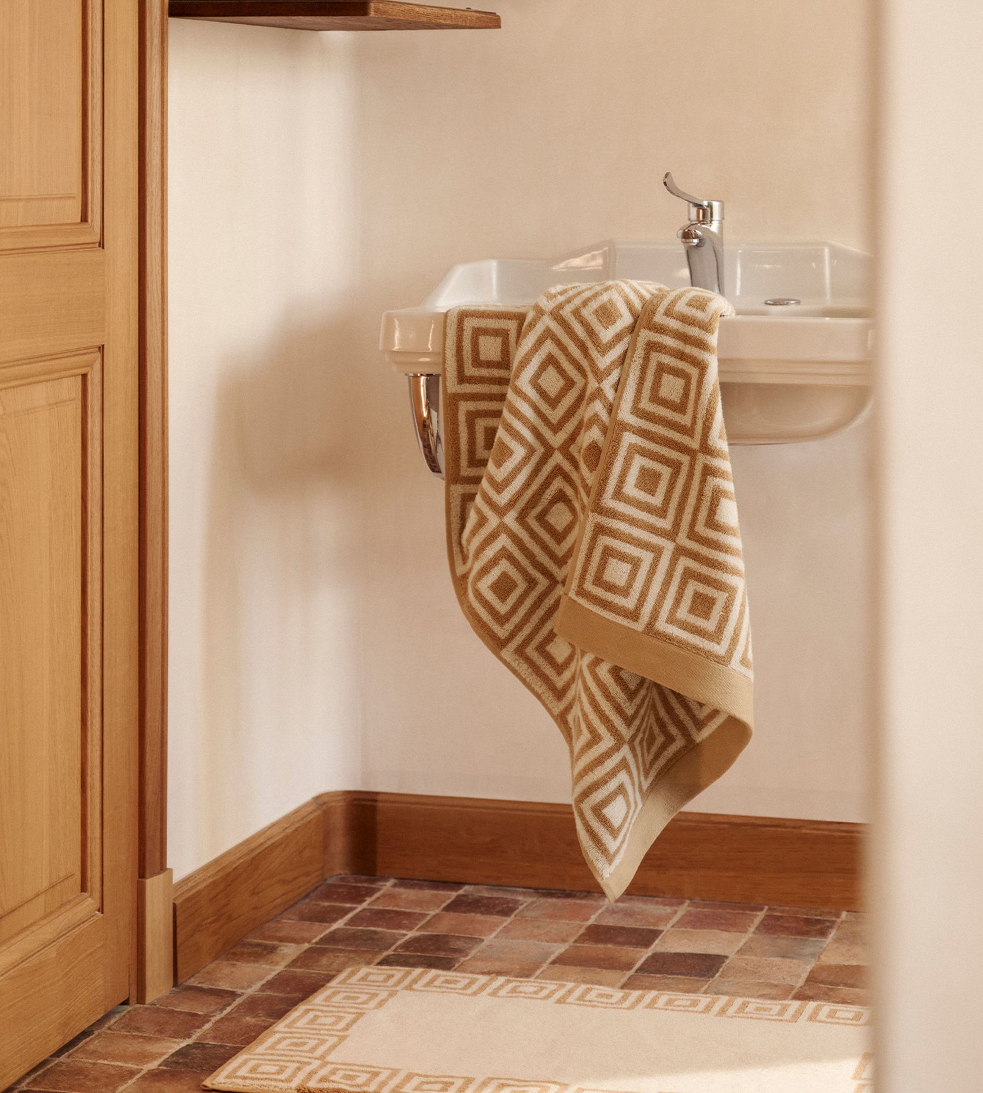 Salle de bain avec une serviette à motifs géométriques suspendue à un lavabo blanc. Tapis assorti sur un sol en carreaux de terre cuite. Murs aux tons neutres et accents en bois.