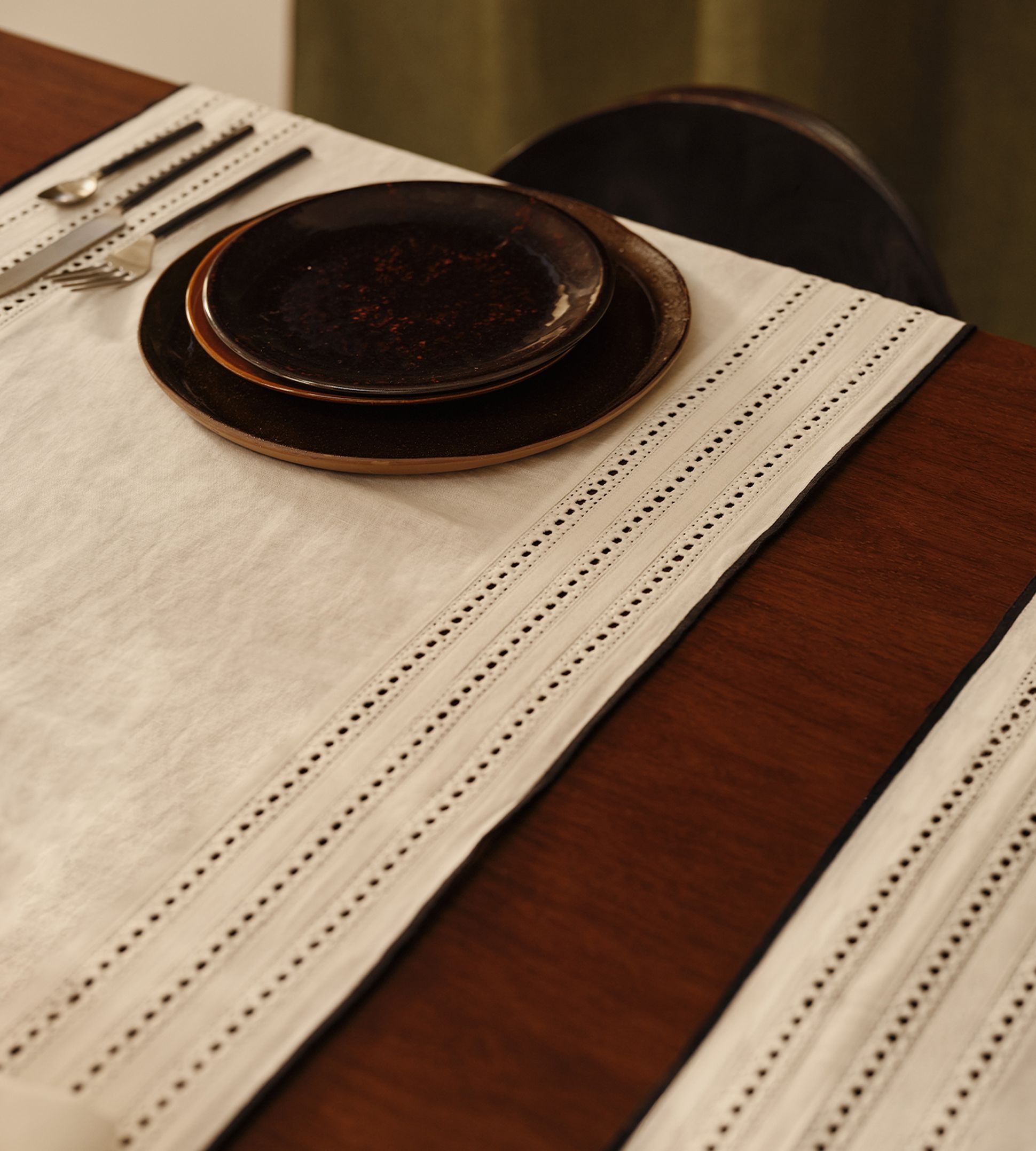 Une table élégamment dressée, avec deux assiettes foncées empilées sur un chemin de table blanc bordé de dentelle, les couverts à gauche et un plateau en bois en dessous.