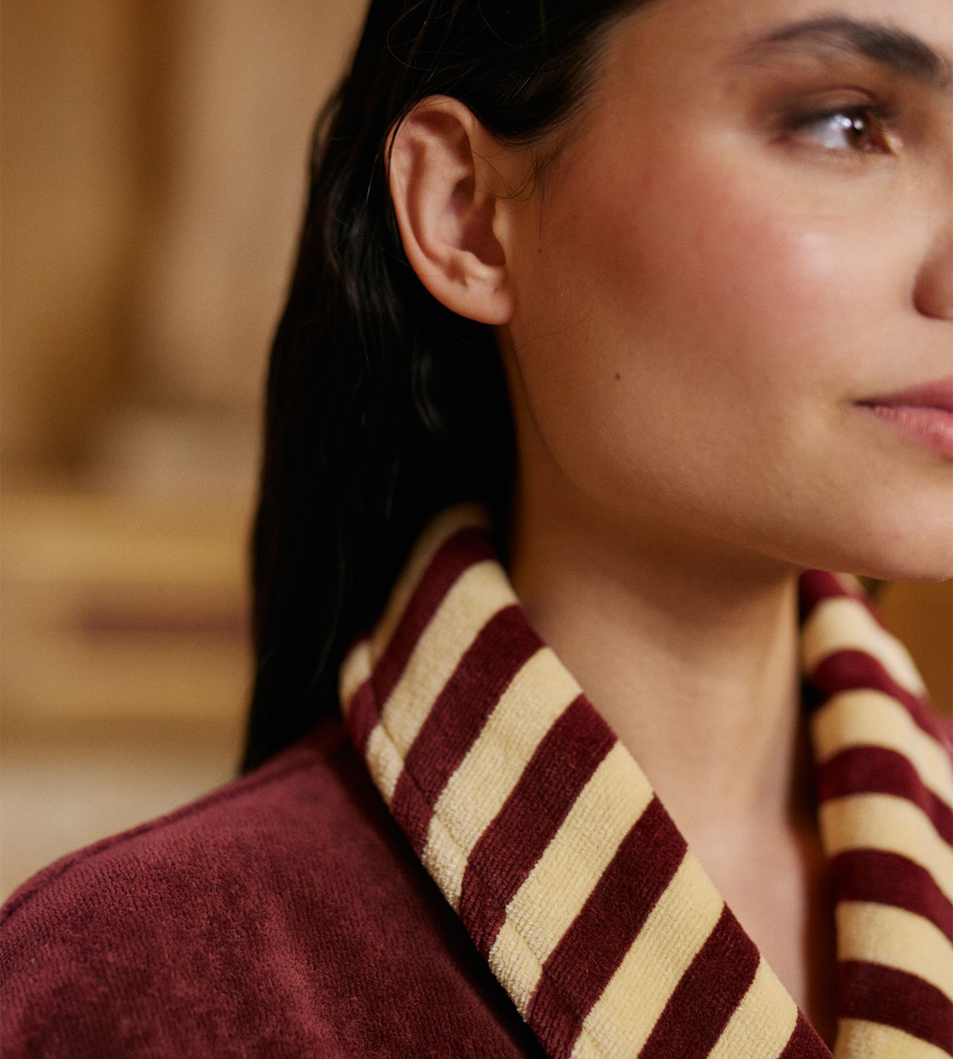 Gros plan sur une femme vêtue d'une chemise bordeaux à col rayé marron et crème, le regard tourné vers la droite.