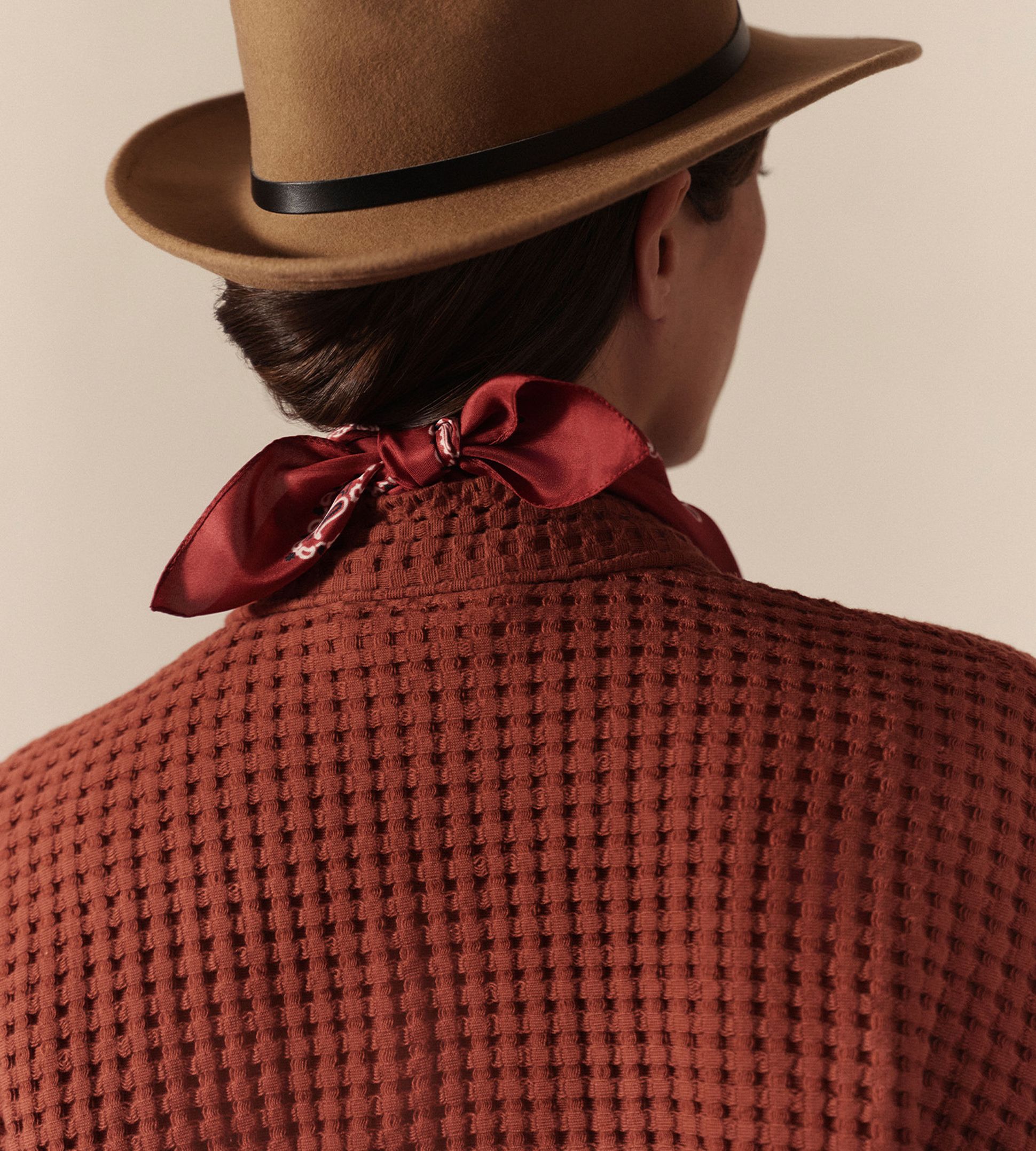 Personne portant un chapeau marron, un haut texturé couleur rouille et une écharpe rouge nouée dans le dos. Elle tourne le dos à la caméra.