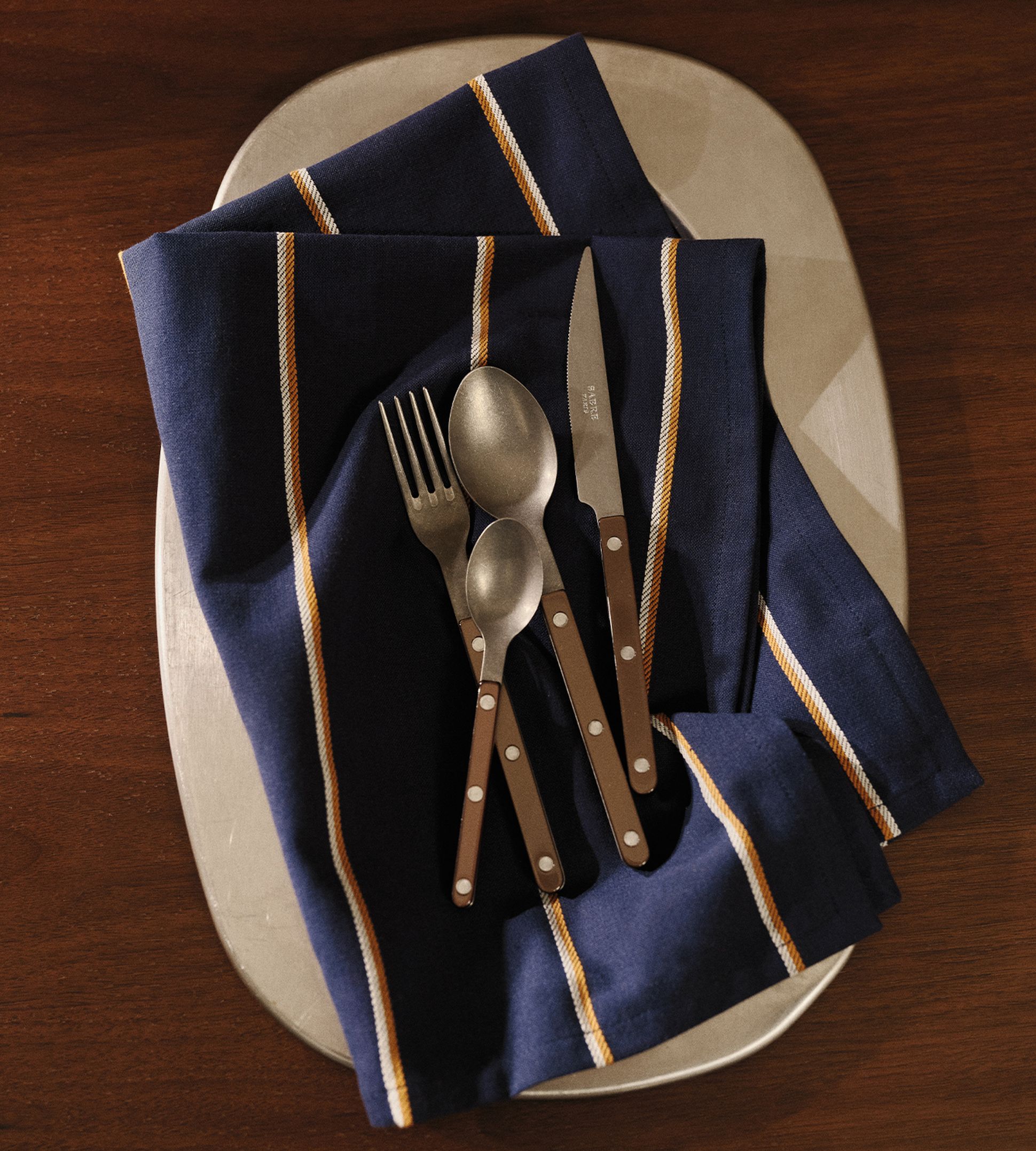 Un service de couverts posé sur une serviette bleue rayée, sur une assiette, le tout disposé sur une table en bois.