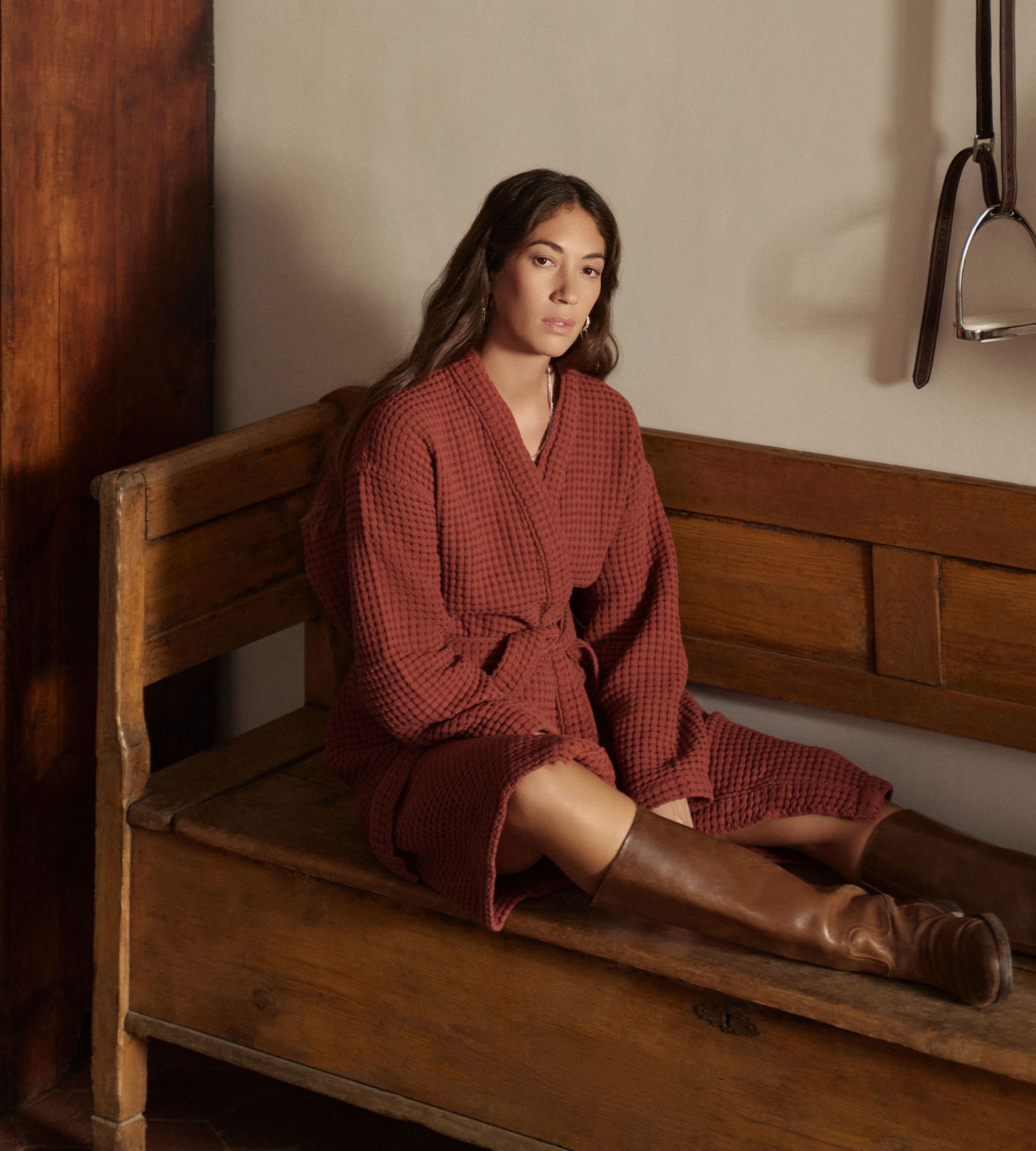 Une femme vêtue d'un peignoir rouge et de bottes marron est assise sur un banc en bois dans une pièce rustique équipée d'une armoire et d'une étagère murale sur laquelle est posée une couverture enroulée.