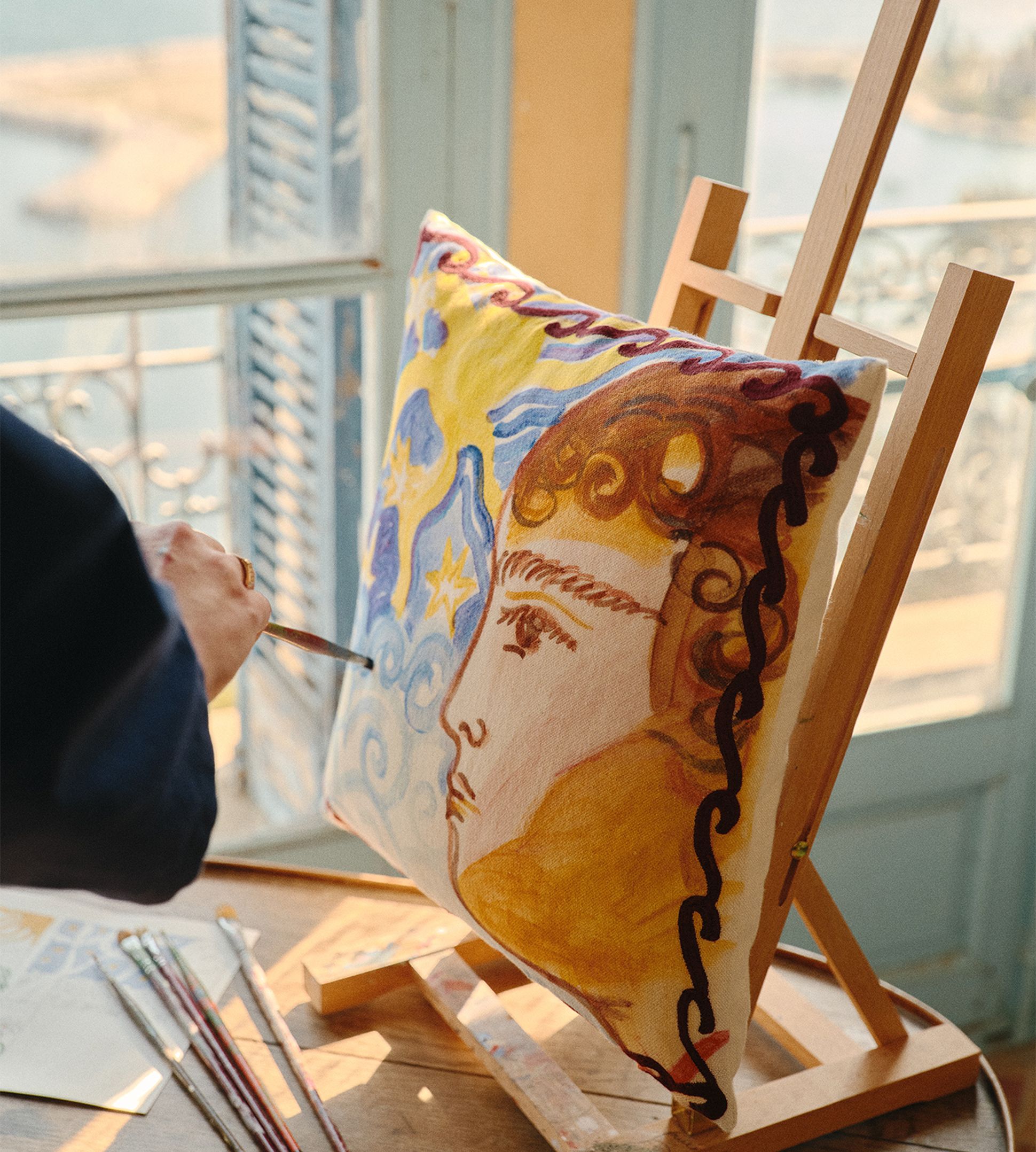 Peindre un portrait haut en couleur sur un coussin, posé sur un chevalet près d'une fenêtre ensoleillée avec vue sur la mer.