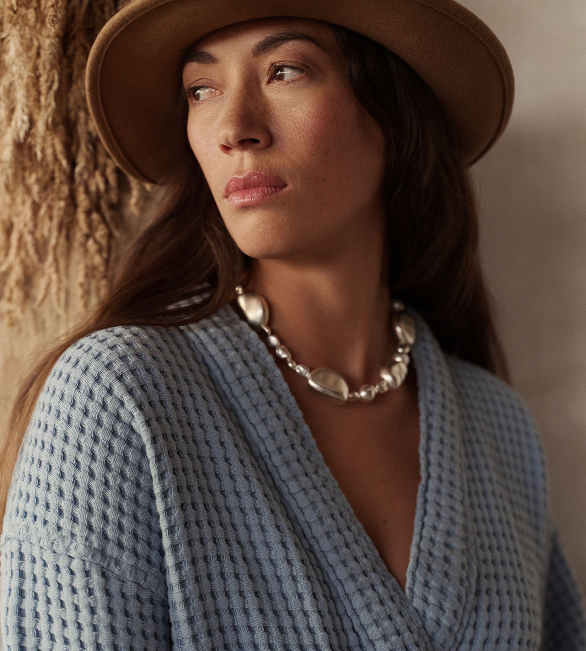 Femme portant un chapeau beige, un collier épais et un pull bleu texturé, regardant pensivement sur le côté. Arrière-plan neutre.