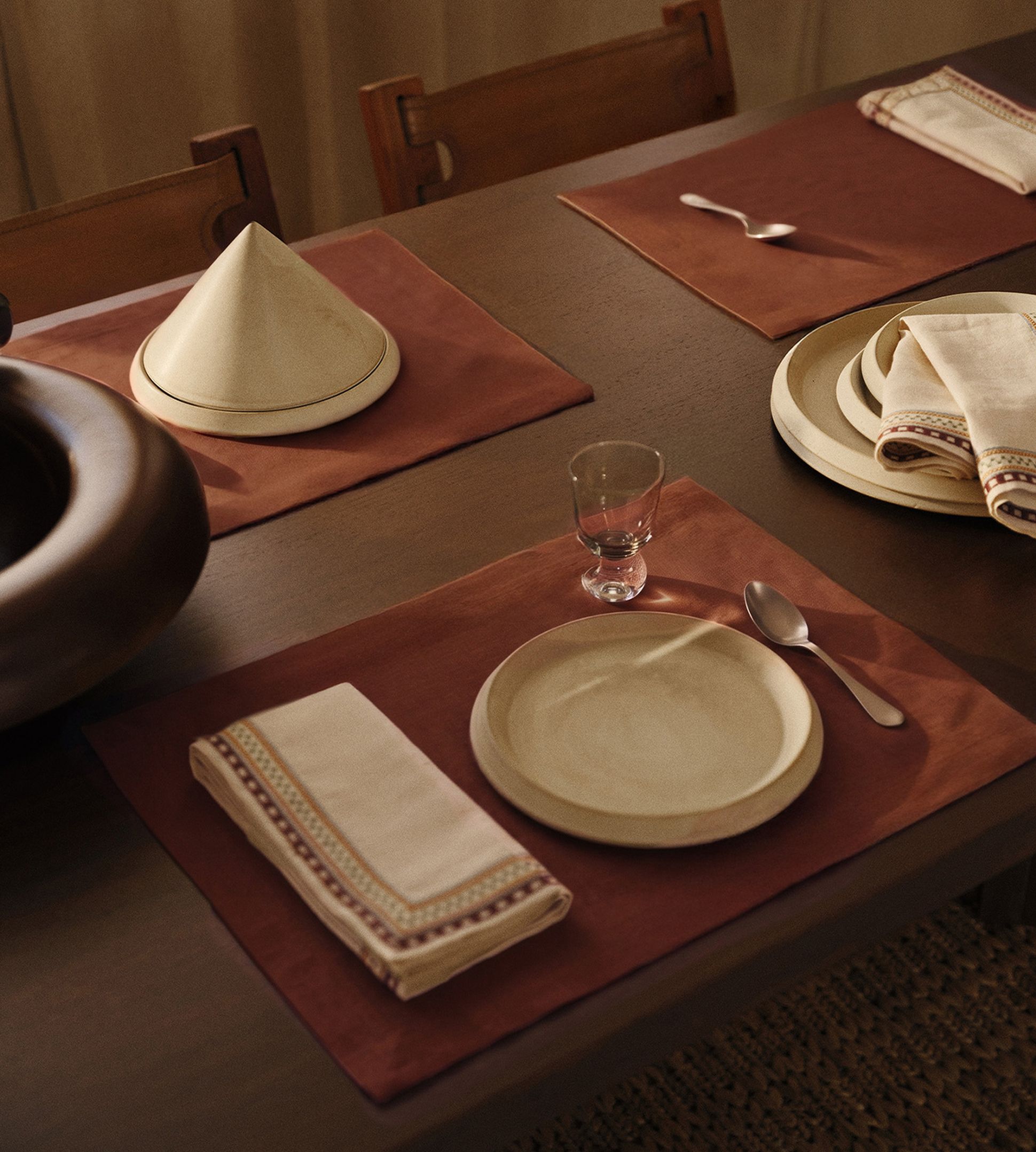 Une table élégamment dressée avec des assiettes beiges, une cuillère, un verre, une serviette pliée et un tajine posés sur des sets de table marron.