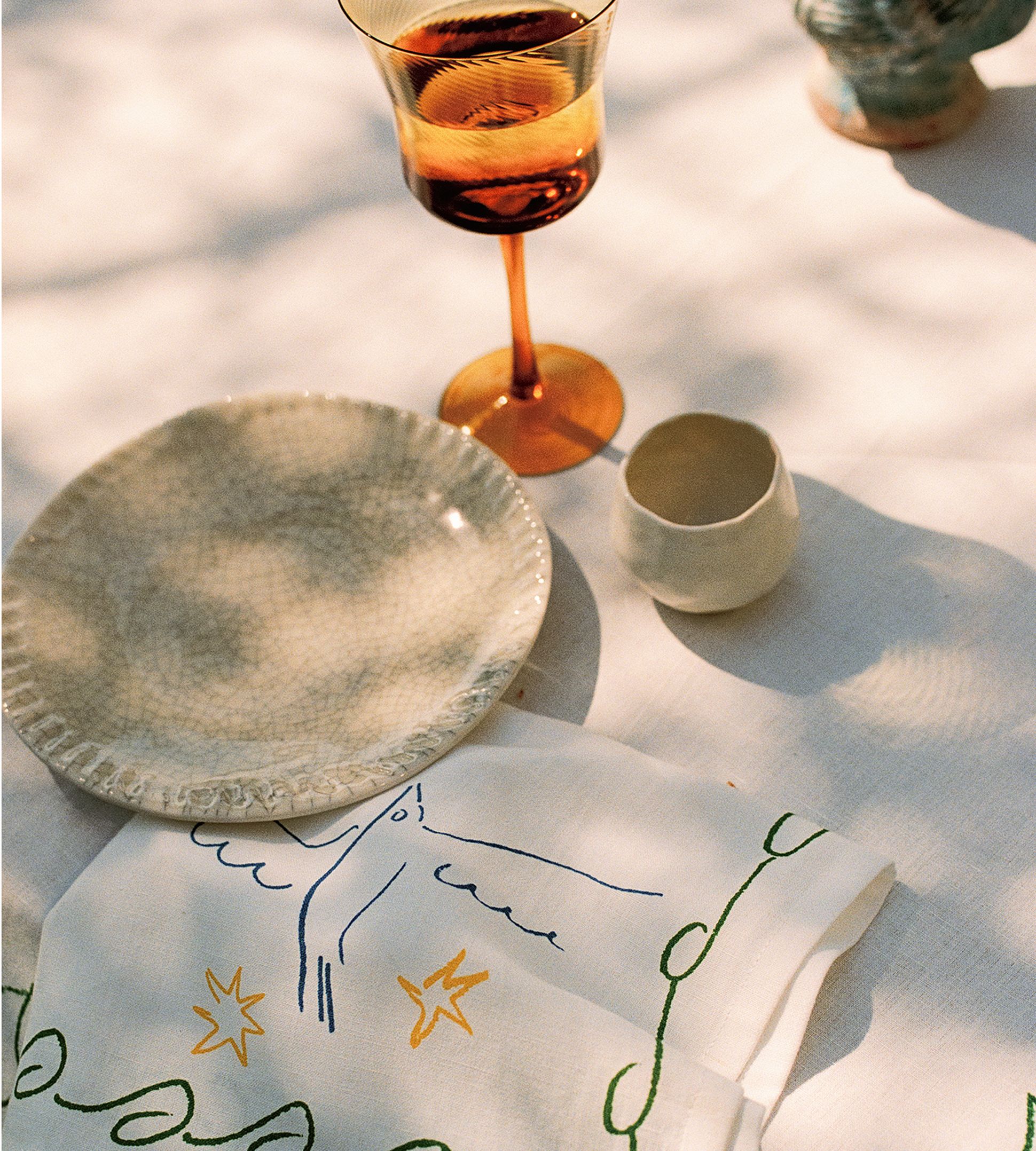 Une table dressée avec une assiette texturée, un verre orange, une petite tasse blanche et une serviette ornée d'oiseaux et d'étoiles, le tout sur une nappe baignée de soleil.