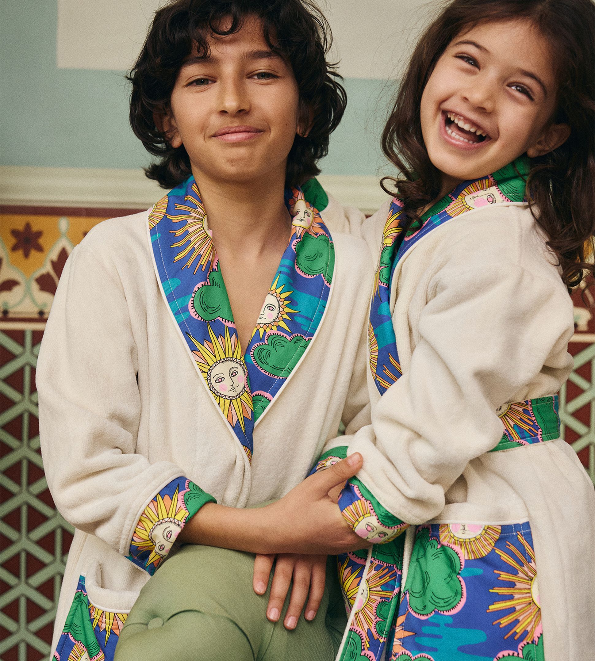 Deux enfants vêtus de robes assorties ornées de motifs solaires colorés sourient joyeusement, enlacés dans un décor chaleureux.