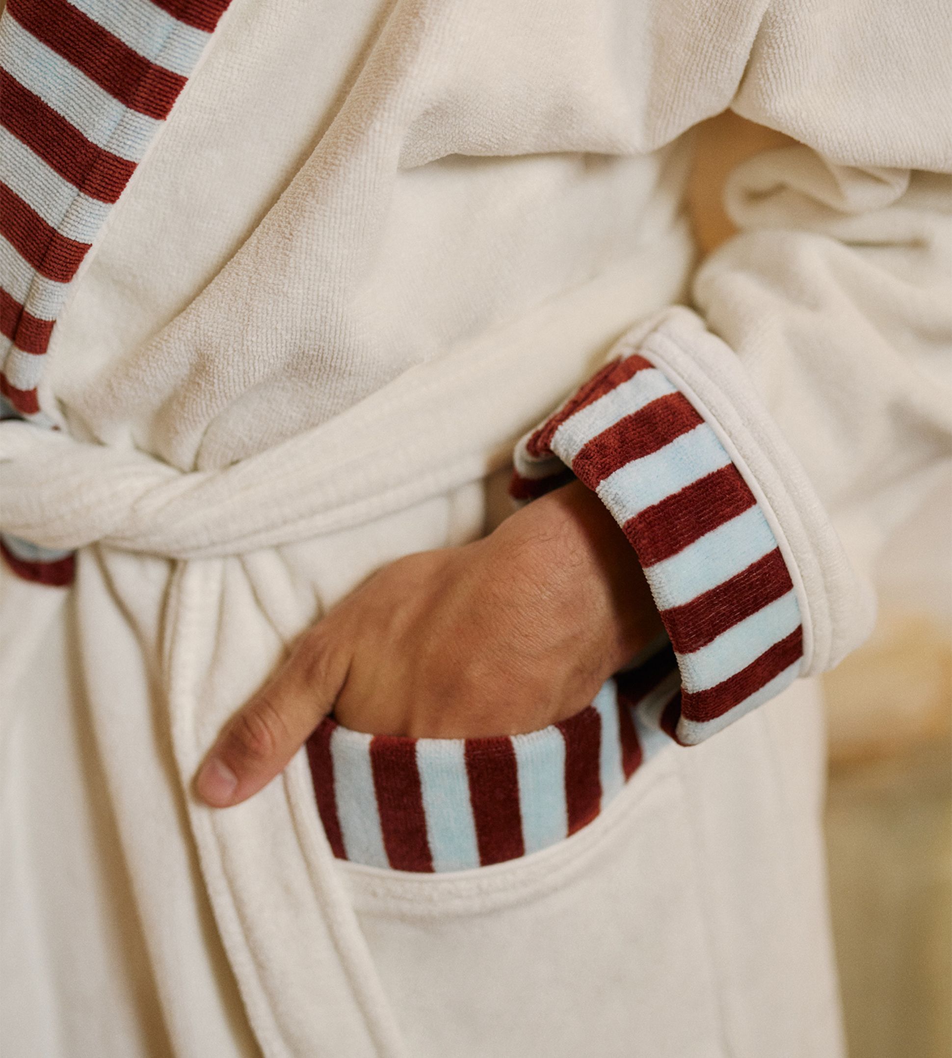 Une personne vêtue d'un peignoir crème aux poignets rayés de rouge et de bleu, la main dans la poche.